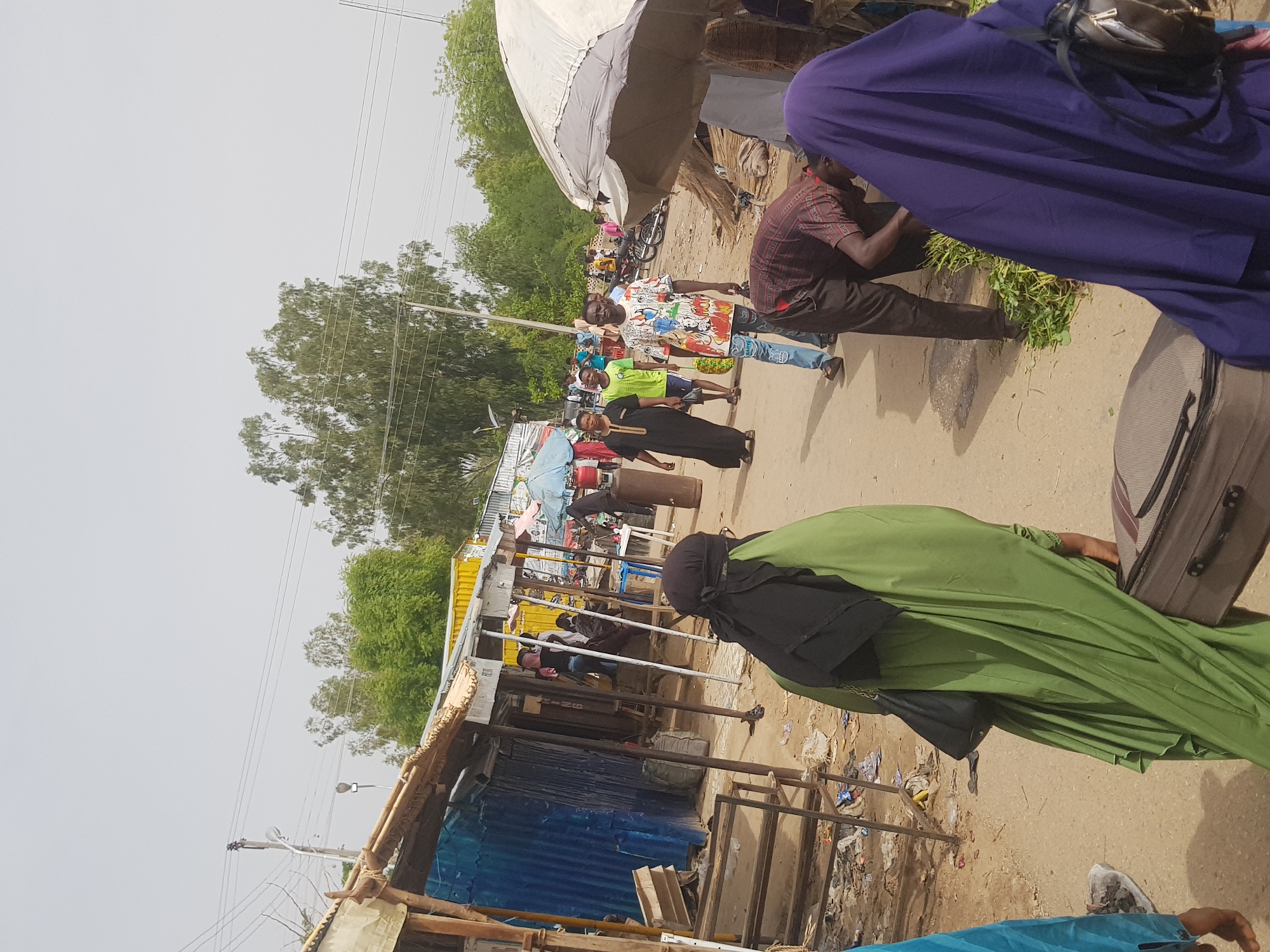 The image depicts an outdoor market scene in Dabawa, Nigeria. The market stretches along a dirt road. Several people are visible: individuals in traditional Islamic clothing, including a woman in a green robe and black head covering, and another in a purple robe. Further down the road, there is a mix of men and women, with one man wearing a cartoon print shirt. Some are standing and walking, others are vendors. The market stalls are constructed with a variety of materials, including corrugated metal and wooden supports. Overhead, electrical wires run parallel to the road. The environment is dusty, and litter is visible near the stalls.