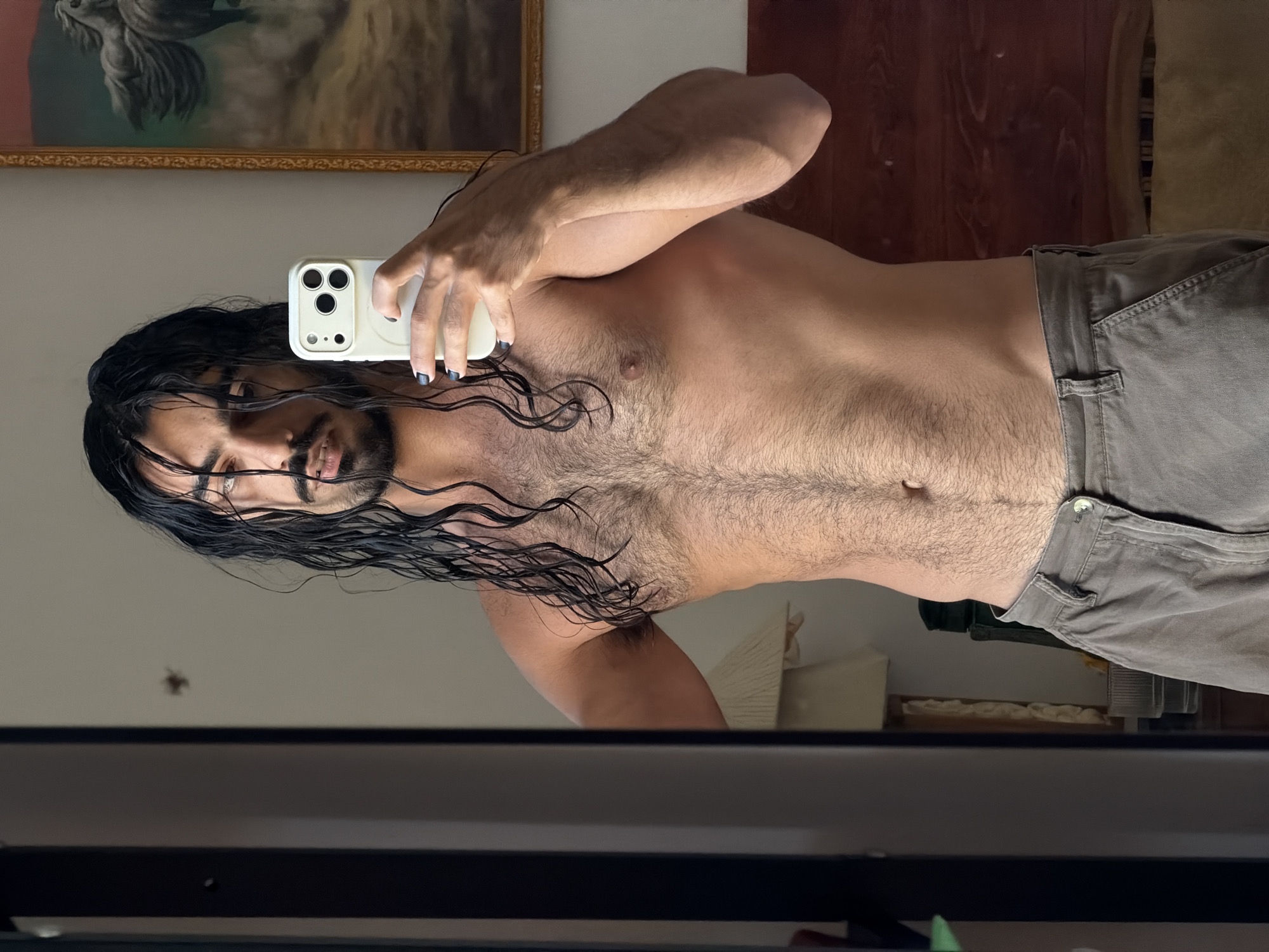 A shirtless man with long, dark, wet hair is taking a mirror selfie. He has a muscular build and is wearing grey pants. His fingernails are painted black. In the background, a painting of horses hangs on the wall. A lamp and other furniture are visible in the reflection. The scene appears to be indoors, likely in a bedroom. The image does not contain any information about a specific location in North Kuta, Indonesia.