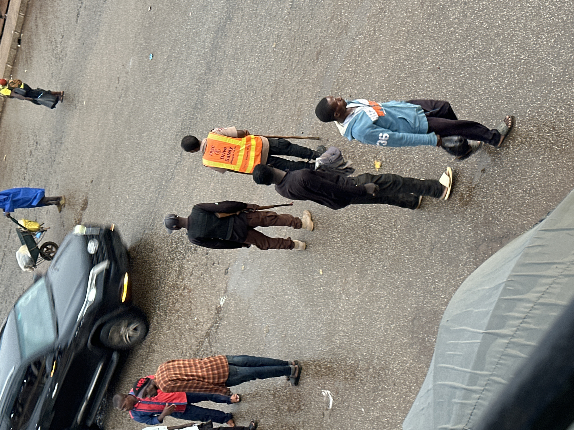 The image shows a street scene in Bukuru, Nigeria, with several people and a black car. The people are standing or sitting on the ground, and some appear to be resting or waiting. One person is wearing an orange reflective vest with the text "FRSC Drive Safely" visible. Another person is wearing a blue hoodie with the number "36" on it. The car is parked on the side of the road, and a cart with yellow bags is visible behind it. The ground is wet, suggesting it might have rained recently. The overall atmosphere appears to be a typical day on a Nigerian street, with people going about their daily activities. The lighting suggests it is daytime.