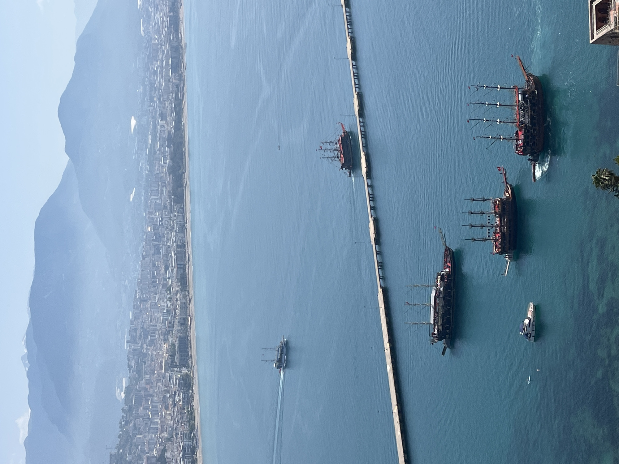 The image captures a high-angle view of the coastline of Alanya, Turkey. Mountains are visible in the background, partially obscured by haze. A city stretches along the coast at the base of the mountains. The sea is a prominent feature, displaying a gradient of blue hues.
A long, concrete pier extends into the sea. Several boats, including stylized "pirate ships" with multiple masts, are visible in the water, some close to the pier. One smaller, modern white boat is also present. There is no visible human presence.