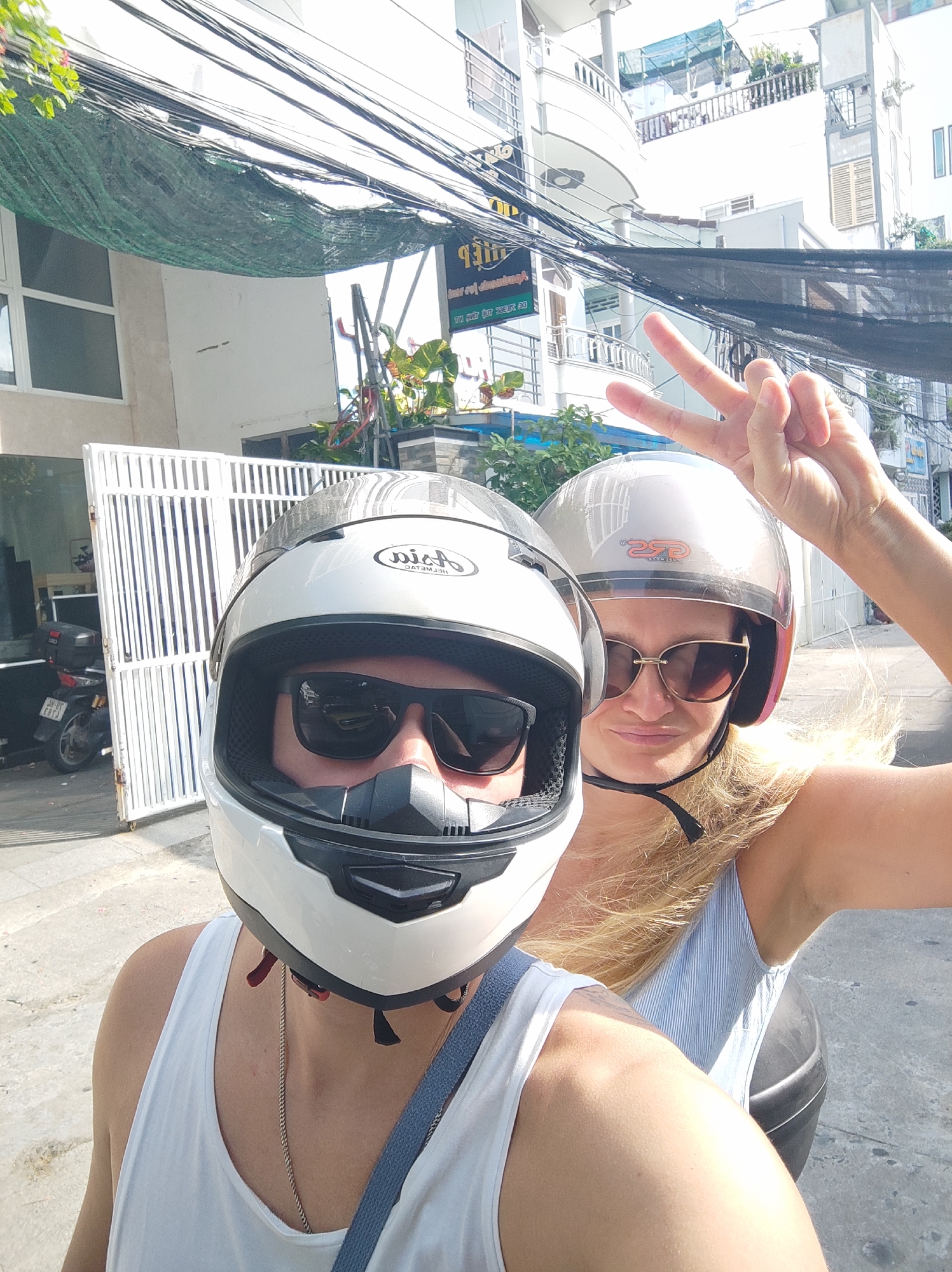 In a street scene in Nha Trang Ward, Vietnam, two individuals are captured in a selfie. Both are wearing helmets, with the person in the foreground wearing a white full-face helmet and dark sunglasses, and the person behind wearing a silver helmet and sunglasses. The individual in the background has blonde hair and is making a peace sign with their right hand. They are both likely on a motorbike, as indicated by the presence of a black seat or storage box visible behind them. The environment features a white metal gate, a parked motorbike with a license plate that appears to be in Vietnamese, and buildings with balconies and wiring overhead. A green tarp is stretched across some of the overhead wires. The lighting suggests it is daytime.