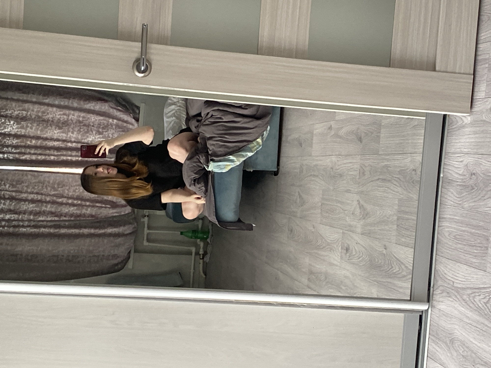 An indoor mirror selfie captures a young woman in what appears to be a bedroom or dressing area in Chaikovsky, Russia. The woman, with long, straight brown hair, is wearing a dark, short-sleeved top. She is positioned in front of a large mirror, holding a dark-colored smartphone with both hands to take the picture. Her legs are bent, suggesting she is squatting or sitting.
The mirror forms part of a light wood-framed sliding door, likely a wardrobe or closet, and features a horizontal metal handle at the top. Reflected in the mirror, behind the woman, are light-colored walls and a patterned curtain to her left. To her right, there is a piece of furniture, possibly a bed or couch, covered with dark blankets and clothing. A green bottle is visible on the light wood-patterned floor near the furniture, along with what appears to be a radiator or heating pipes on the wall. The lighting is ambient indoor light, and no strong shadows are present. No visible text is discernible in the image.