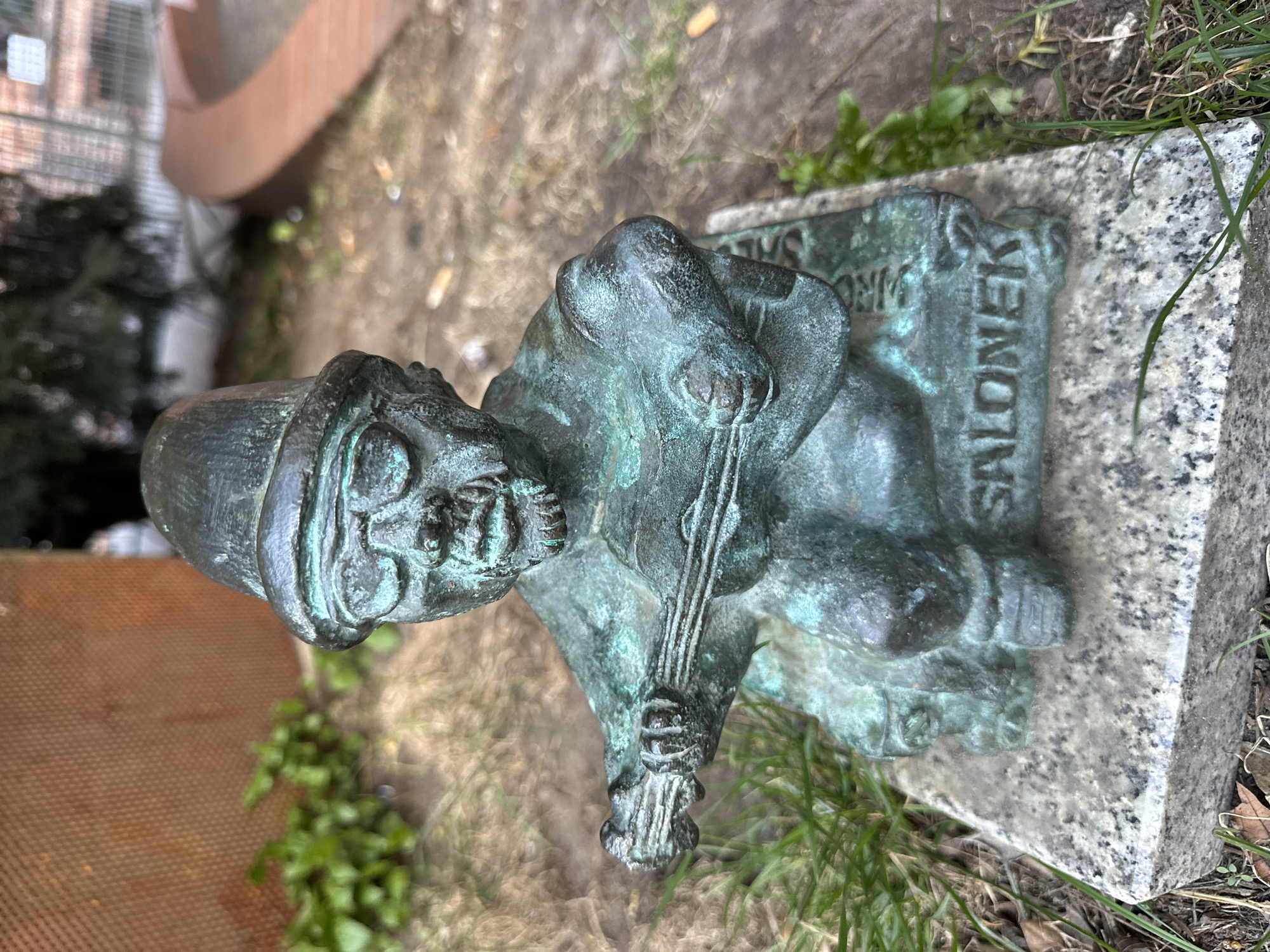 The image shows a bronze sculpture of a dwarf playing a cello, located in Wrocław, Poland. The dwarf is wearing a hat and seated on a stone block that has the name "SALONEK" inscribed on the side. The sculpture is positioned on the ground amidst grass and some loose soil. Behind the sculpture, part of a building and a curved, brown structure are visible.