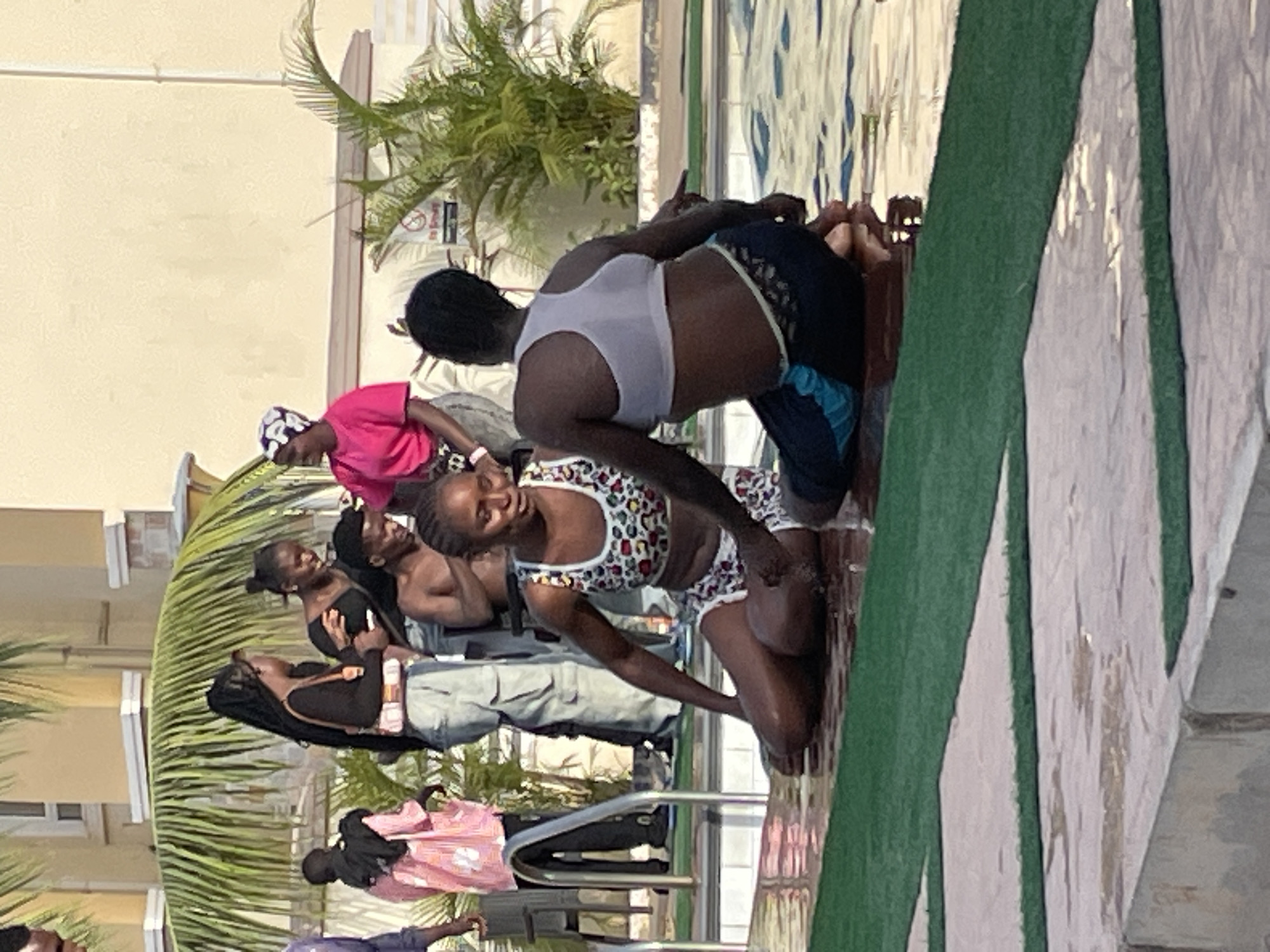 A group of people are gathered at a pool's edge in what appears to be a recreational setting. The location is Barakin Lerawi, Nigeria. Two individuals are physically close, one in a white tank top and blue shorts and the other in a patterned bathing suit. They are at the edge of a pool, close to the water. Additional people are standing nearby, one wearing a pink top and patterned head covering, another wearing black, and others in casual attire. A palm tree is visible, as is part of a building.