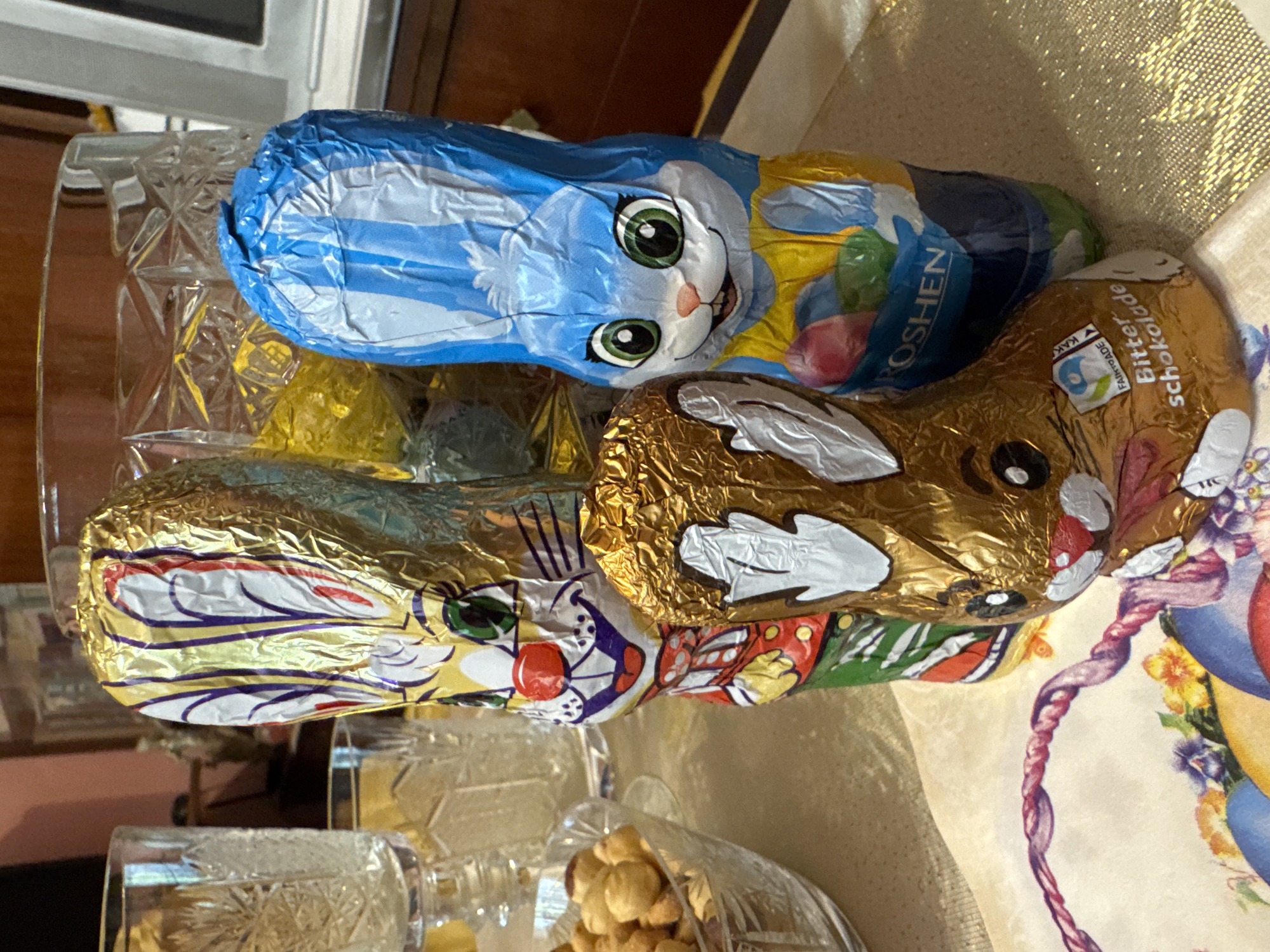 The image shows a close-up of three foil-wrapped chocolate bunnies on a table. The bunnies appear to be sitting on or near a crystal bowl containing nuts. The background is blurred, but suggests an indoor setting with a patterned tablecloth. The bunny on top has a blue and white wrapper. The bunny beneath it has a gold wrapper. The third bunny also has a gold wrapper with white and purple markings. The text "Bitterschokolade" is visible on the lower bunny. A label on the same bunny indicates fair trade. There is no explicit indication of the location being Plovdiv, Bulgaria.
