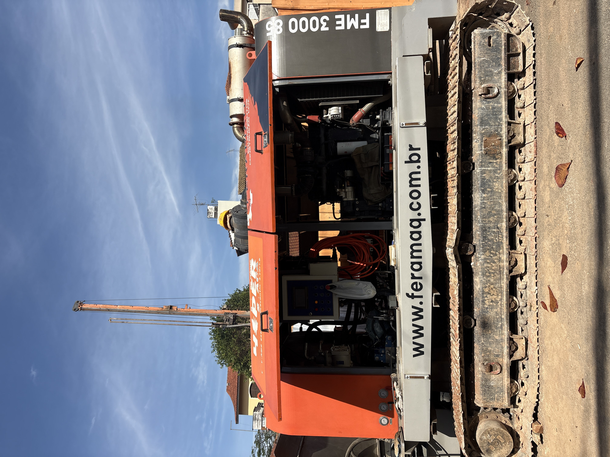 This image depicts a large, orange and grey drilling rig, possibly a FME 3000 85 model, parked on a dirt road in a city. The rig has caterpillar tracks and an open engine compartment revealing various mechanical components. A yellow hard hat is visible on a person obscured by the machinery, suggesting active construction work. The rig bears the website "www.feramaq.com.br" and the text "FME 3000 85" vertically on its side. In the background, there are residential buildings and trees under a clear blue sky with wispy clouds. The scene appears to be a daytime shot in a Brazilian city, likely São Paulo given the ".com.br" domain, with a focus on heavy construction equipment.