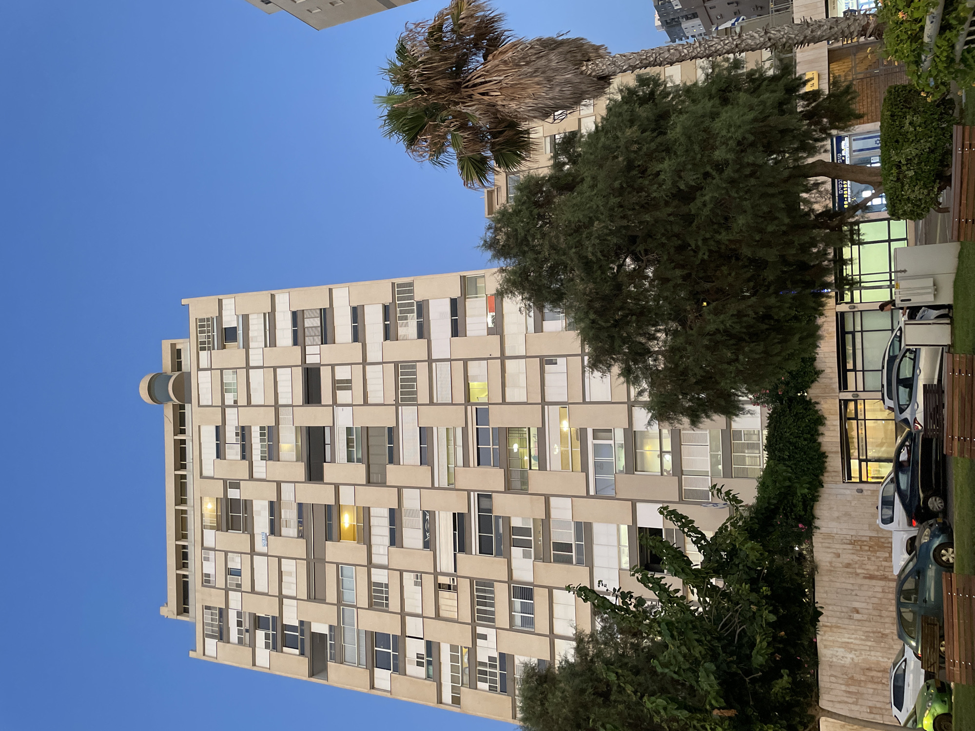 The image shows a tall, beige apartment building in Netanya, Israel, with a patterned facade featuring numerous windows and balconies. The sky above is clear and blue, indicating it's likely early evening. In the foreground, there are several cars parked along the street. Trees and a palm tree are visible in front of the building, and some benches are placed on a grassy area. The building's windows are a mix of open and closed, with some lights on inside, suggesting residents are present. The overall impression is of a residential urban environment in the late afternoon or early evening.