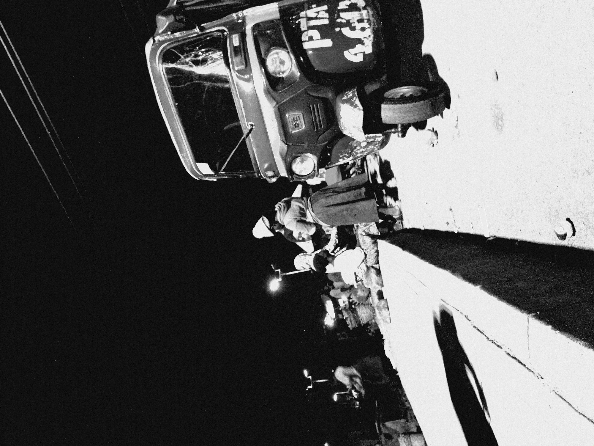 At night in Bukuru, Nigeria, a bustling marketplace unfolds. A three-wheeled auto-rickshaw, its front damaged and bearing the markings "PTA 4613," is parked on a dusty road. A woman in a white headscarf and a long skirt stands beside it, while another woman in a headscarf and a long, dark skirt is seated nearby. In the background, more figures are visible amidst market stalls and illuminated by streetlights, suggesting ongoing commerce and activity. The scene is characterized by strong contrasts between light and shadow, typical of nighttime street photography in a developing urban environment.
