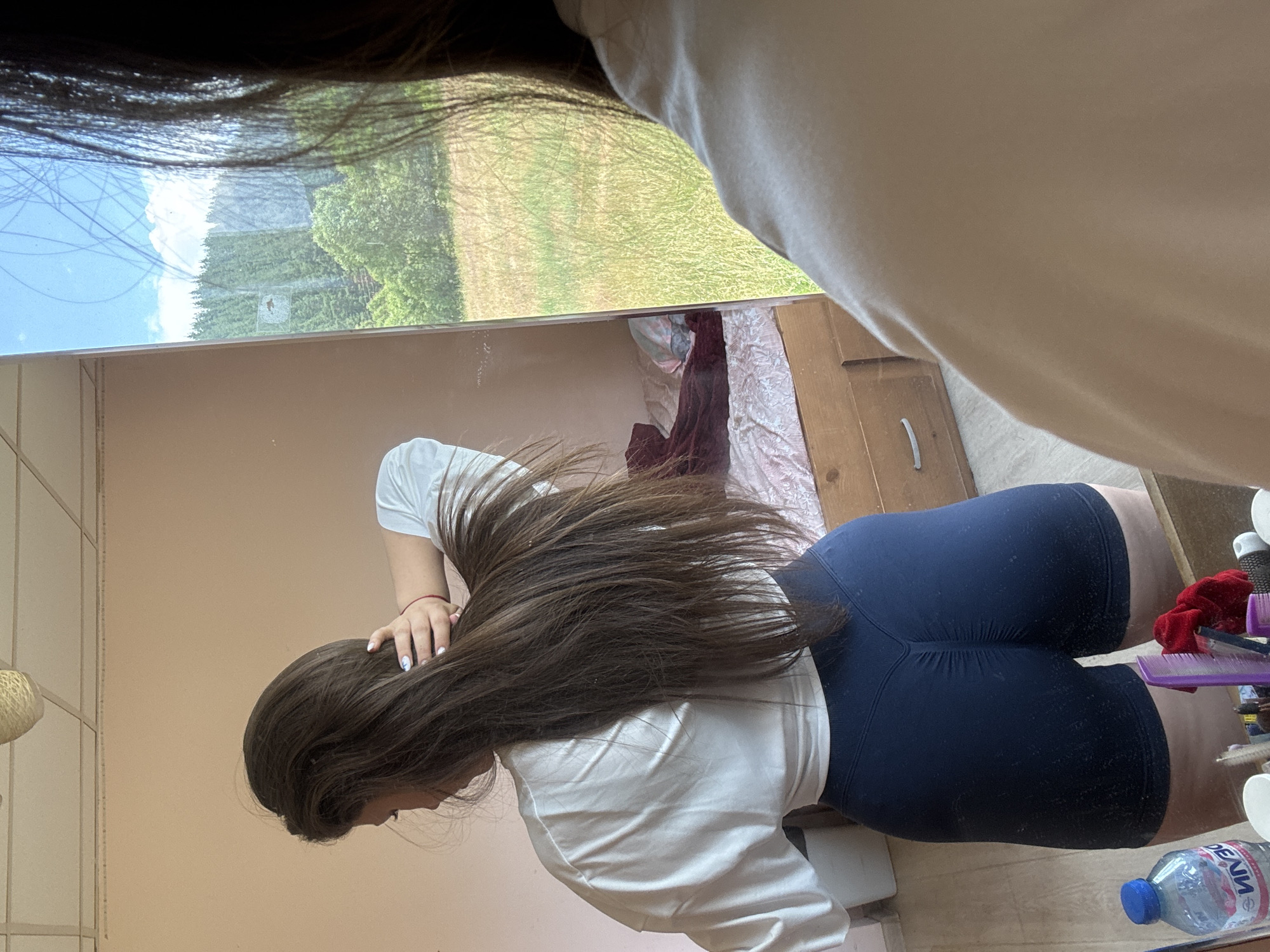A young woman with long, dark hair is standing in a room, facing away from the camera and looking into a mirror. She is wearing a white crop top and dark blue shorts. Her right hand is raised to her head, touching her hair. In the mirror's reflection, a bed with bedding is visible in the background, along with a dresser with drawers. The room appears to be a bedroom. Outside the window, reflected in the mirror, is a landscape with green trees and a field under a blue sky with clouds. This scene is located in the village of Prodanovtsi, Bulgaria.