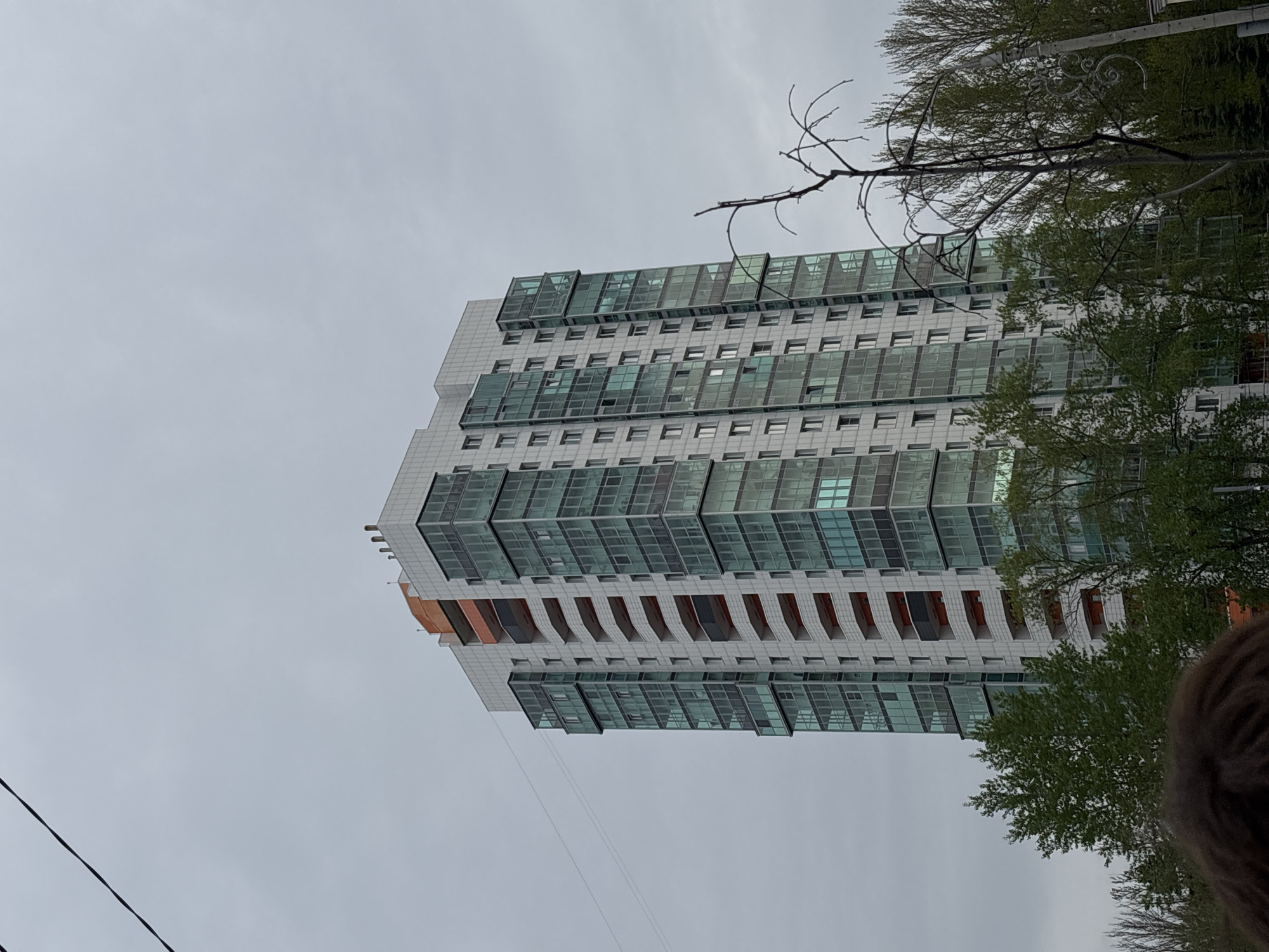 The image shows a tall, modern building in Perm, Russia. The building is predominantly white with multiple floors, each featuring numerous windows. Some floors have distinct architectural elements like recessed balconies. The sky is overcast. Trees are visible on the right side of the frame, partially obscuring the building. Power lines are visible in the lower-left corner. A portion of a person with dreadlocks is visible at the bottom right of the image.