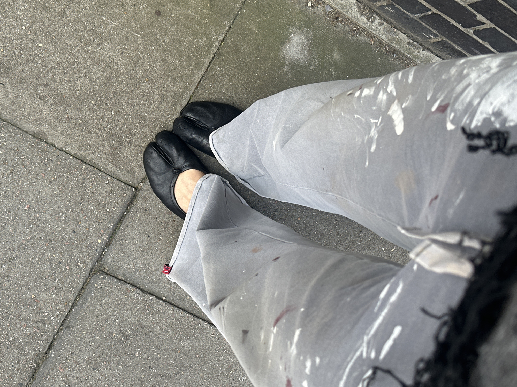 An overhead shot shows a person standing on a sidewalk composed of grey rectangular pavers. The person is wearing grey trousers with splatters of white and red paint. The trousers are worn with black Tabi shoes. The location is the city of London, United Kingdom.