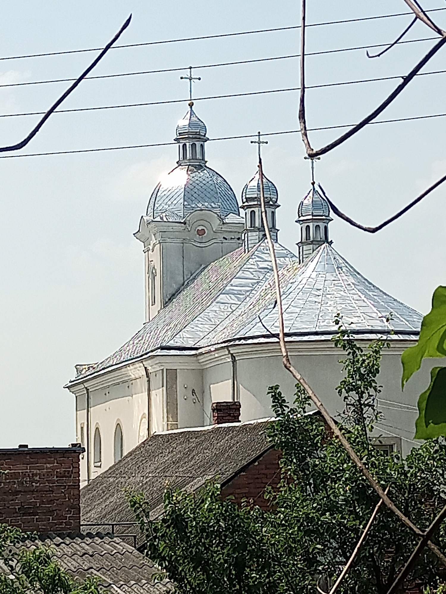 The image shows a white church with silver domes and crosses, partially obscured by tree branches and power lines. The church appears to be in the city of Yampil, Ukraine. The foreground consists of rooftops, vegetation, and the aforementioned branches and wires. The sky is clear and light blue.