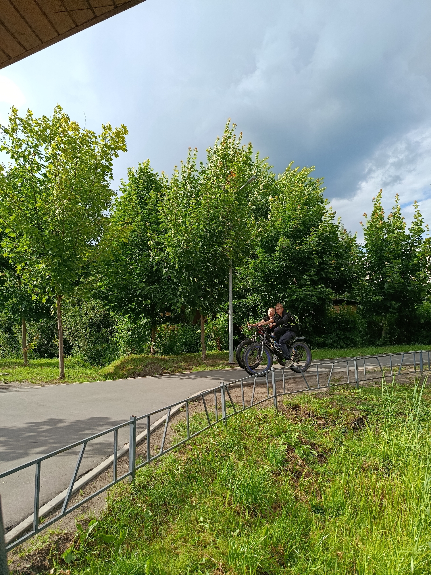 Two individuals are riding a single fat tire bicycle on a paved path. The person in the back is steering, while the person in front is looking back. The bicycle is black with blue accents on the wheels. They are surrounded by lush green trees and grass, with a cloudy sky overhead. A metal fence runs along the right side of the path. The scene appears to be in a park or recreational area within the city of Vereya, Russia.