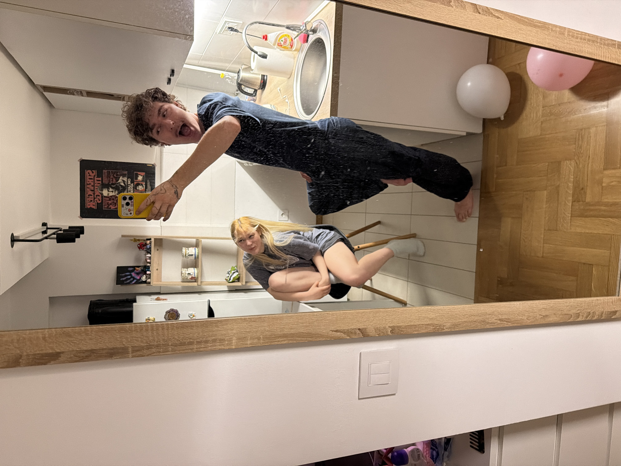 The image, taken in what appears to be a small apartment kitchen in Vracar Urban Municipality, Serbia, captures a mirror selfie of two individuals. On the left, a woman with blonde hair is seated on a dark chair, leaning back with her legs tucked in, wearing a grey t-shirt and shorts with white socks. She is looking towards the mirror. On the right, reflected in the mirror, a young man with curly brown hair is taking the photo with a yellow smartphone. He is wearing a dark blue t-shirt and dark pants, striking a dynamic pose with his mouth open in an exclamatory gesture.
The environment behind them includes kitchen cabinets, a countertop with a sink, a kettle, and a bottle of dish soap. A shelf unit is visible, displaying various items including what appear to be small figurines and decorative jars. The floor is covered in a parquet pattern. In the foreground, reflected on the floor, are two balloons, one white and one pink. A light switch is mounted on the wall to the left of the mirror. The overall impression is of a casual, candid moment captured within a domestic setting.