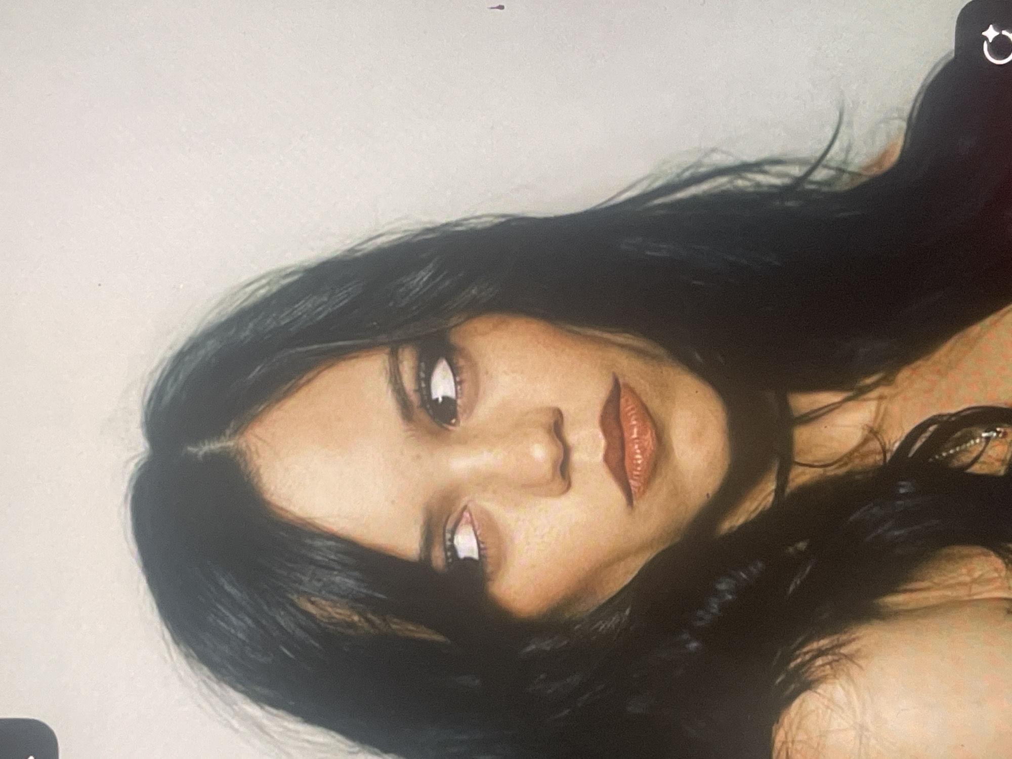 The media file displays a close-up portrait of a young woman with long, dark, wavy hair, oriented sideways (rotated clockwise by 90 degrees). Her head and upper torso are visible against a plain, light off-white background. She has striking dark eyes with prominent reflections, a distinct nose, and full lips accentuated with dark reddish-brown lipstick. Numerous freckles are visible across her nose and cheeks, and her skin has a warm tone.
She appears to be posing for the camera, gazing slightly upwards and to the viewer's right. Her expression is serene and composed. Visible details include a partial view of what appears to be a necklace with a metallic element near her chest. The lighting illuminates her face, creating subtle shadows that define her features.
The image contains digital interface elements: a circular "rotate" icon with a star-like design in the top right corner and a partial dark icon in the bottom left, suggesting this is a screenshot or a photograph of a screen. A small dark smudge is also present near the top center of the background. While the provided context states the location is Poltava, Ukraine, there are no visual cues within the image itself to confirm this. No legible text is present.