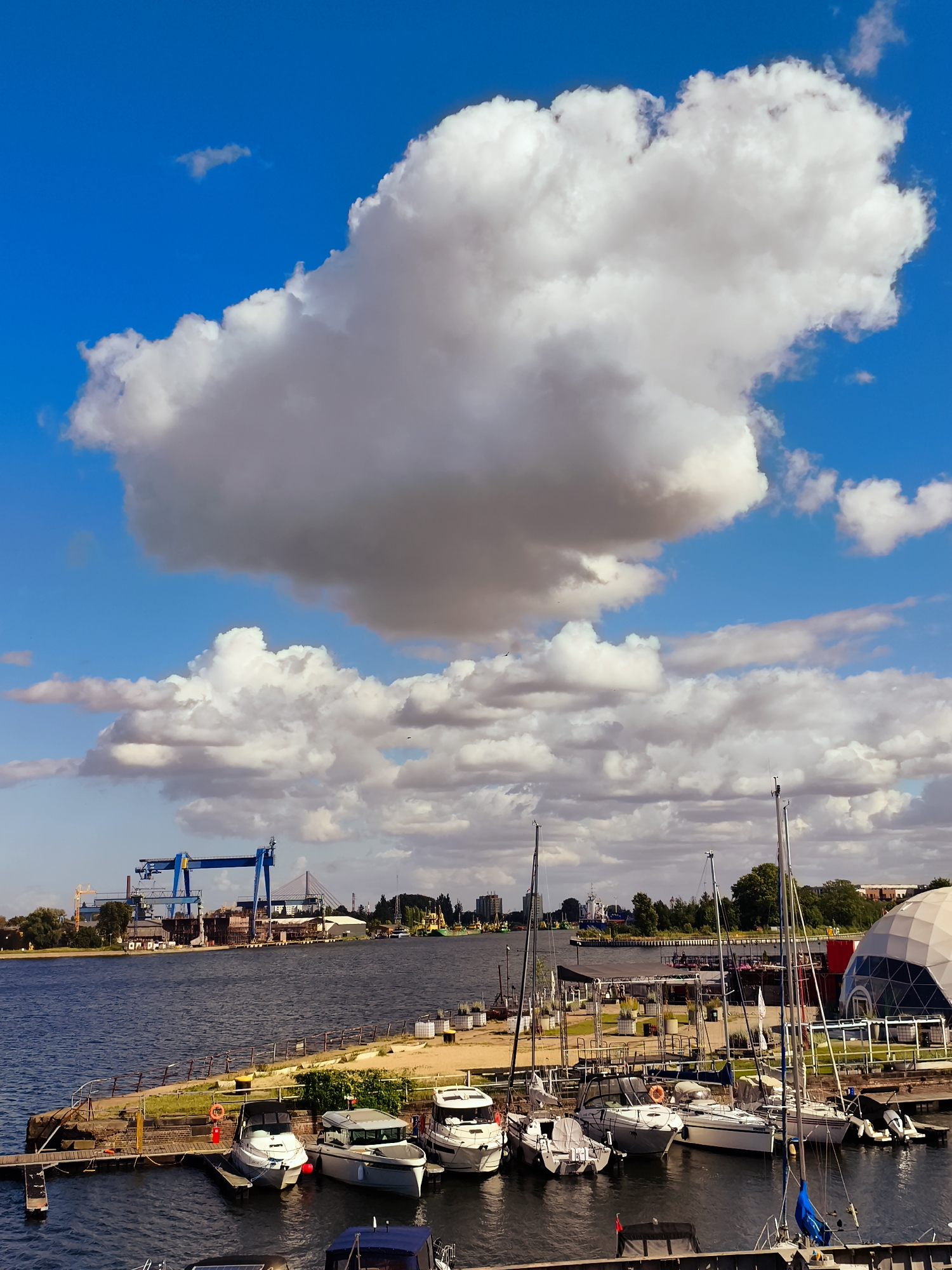 The image depicts a waterfront scene in the city of Gdańsk, Poland, likely within the area of the Gdańsk Shipyard. In the foreground, several yachts and smaller boats are moored at a dock, their masts reaching towards a bright blue sky dotted with large, fluffy white clouds. The water is calm, reflecting the sky and the surrounding structures.
Beyond the docked boats, a promenade or walkway lines the waterfront, with a green strip of vegetation. Further back, across a wide body of water, the industrial landscape of the shipyard is visible. A prominent, large blue gantry crane dominates the skyline, indicative of shipbuilding or repair activities. Other industrial buildings and structures are scattered across the far bank.
To the right of the frame, a distinctive geodesic dome structure, possibly part of a modern building or exhibition space, is partially visible. The overall atmosphere is one of a working harbor combined with recreational boating, under a dynamic, cloudy sky. No people are clearly discernible in the image.