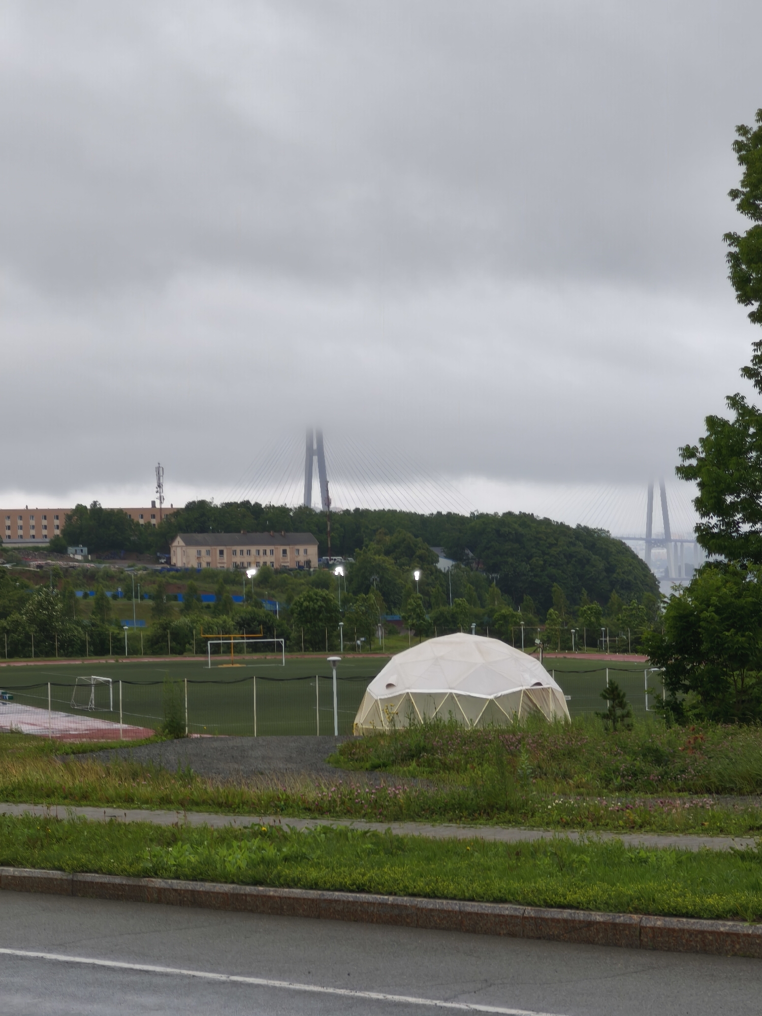 The image depicts a view of a sports complex and infrastructure on Russky Island, Russia, under an overcast sky. In the foreground, a paved road with a white line marking is visible. Adjacent to the road is a strip of green grass and a concrete curb with a granite border. Further back, a pathway leads to a sports field. A white geodesic dome structure is situated on a grassy area to the right of the path, with wildflowers growing around it. The sports field itself features a running track and what appears to be a soccer field with goalposts. In the middle ground, several multi-story buildings, possibly dormitories or residential blocks, are nestled amongst lush green trees. A telecommunications tower stands tall behind one of the buildings. Dominating the background is a large cable-stayed bridge, with its towers partially obscured by mist or clouds. The overall atmosphere is somber due to the cloudy weather. There are no people or animals visible in the frame.