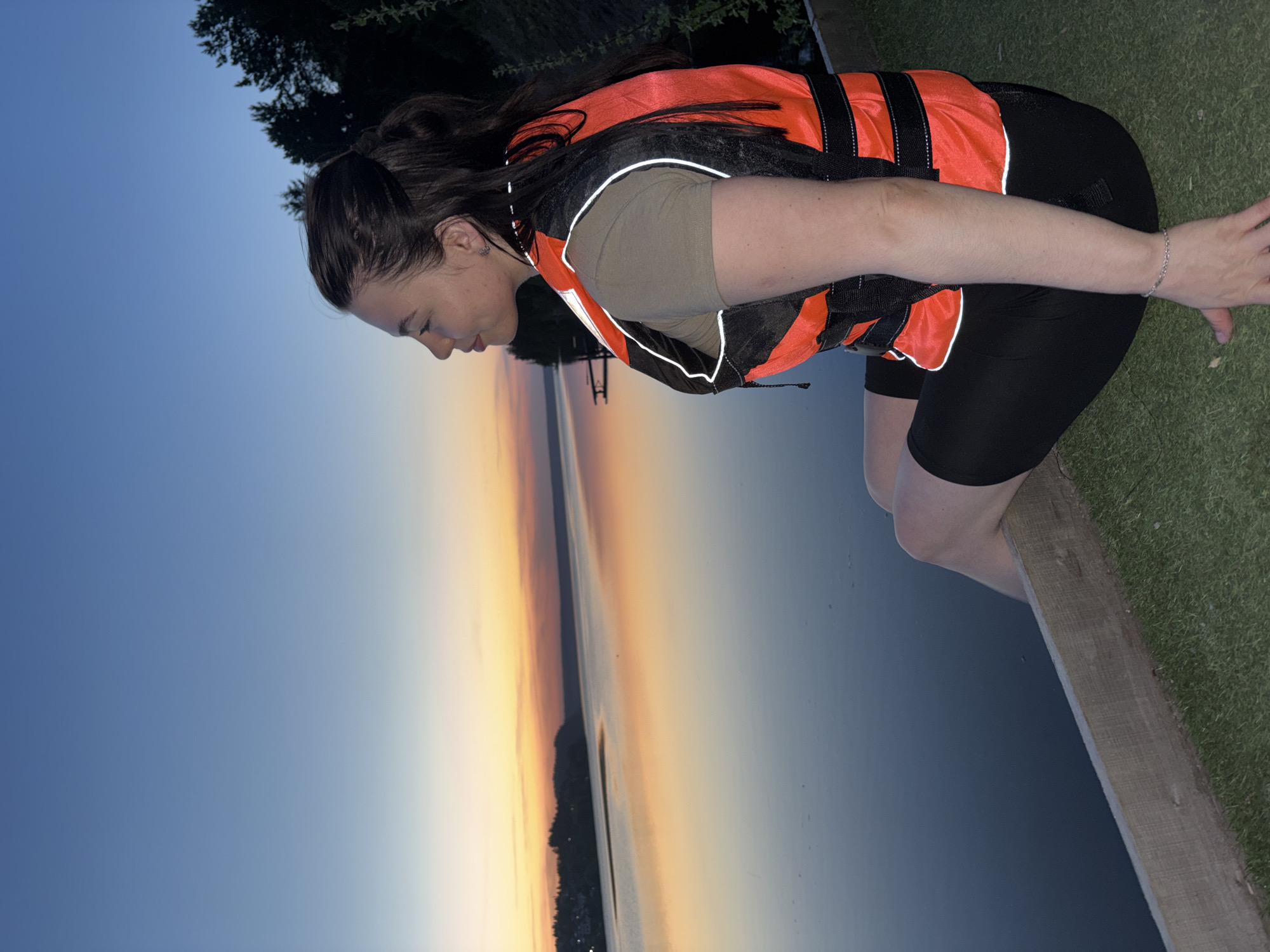 A woman wearing an orange and black life jacket, black shorts, and a green shirt is on a wooden dock at a lake. The background features a sunset sky gradient from light orange to blue over the water. Trees are visible in the upper left. The location is not specified as being Khokhlovka, Russia.