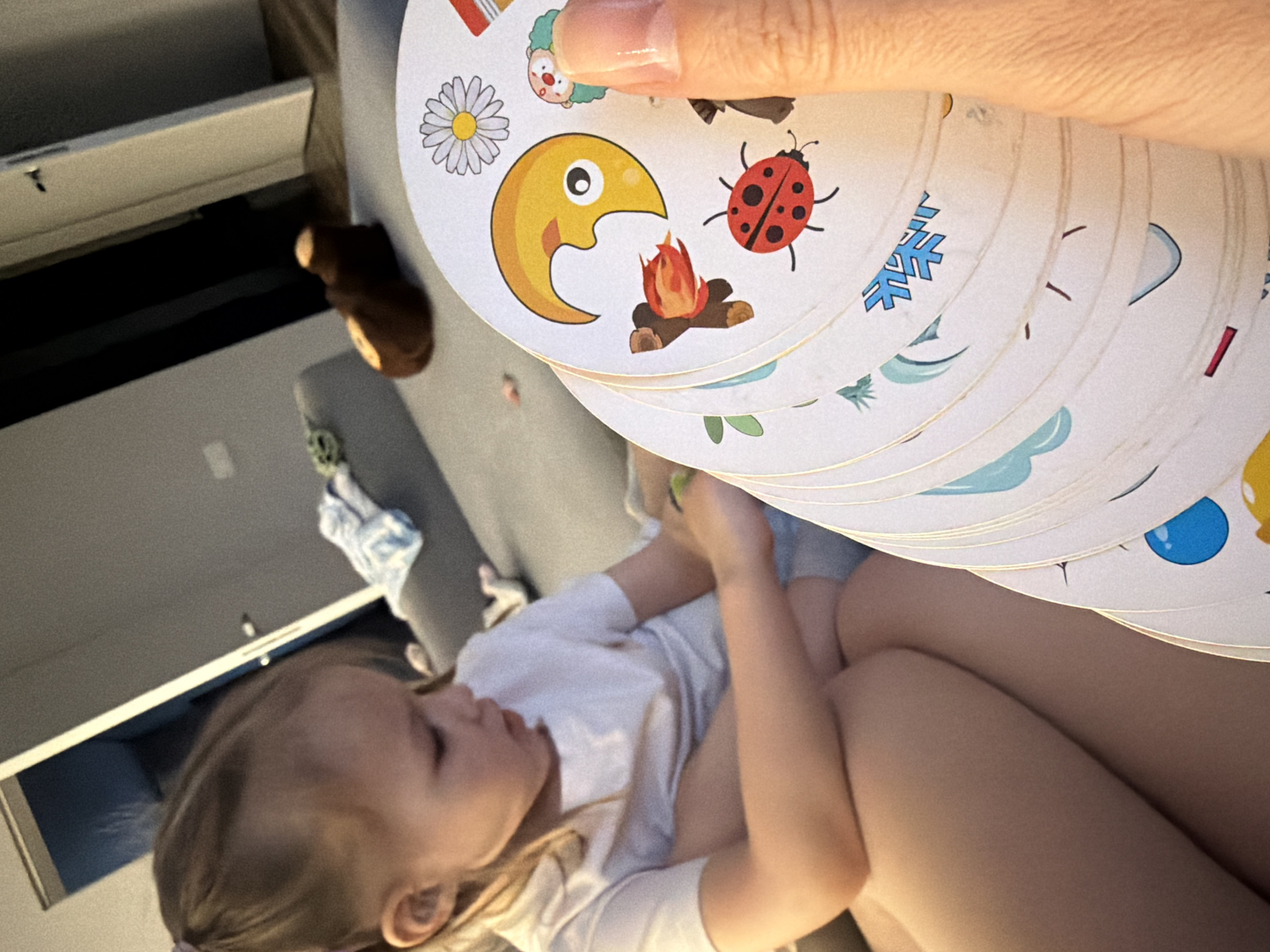 A fair-skinned child is lying on a light-colored upholstered surface, possibly a couch or bed, while a person holds a stack of circular cards with images of a daisy, cartoon figure, ladybug, snowflake, leaf, raindrop, and a yellow crescent moon “eating” a campfire. The child wears a light-colored garment. Visible in the background are elements of a room, including a drawer, and a stuffed toy. The location appears to be a domestic interior but is not definitively identifiable as the city of Chaikovsky, Russia.