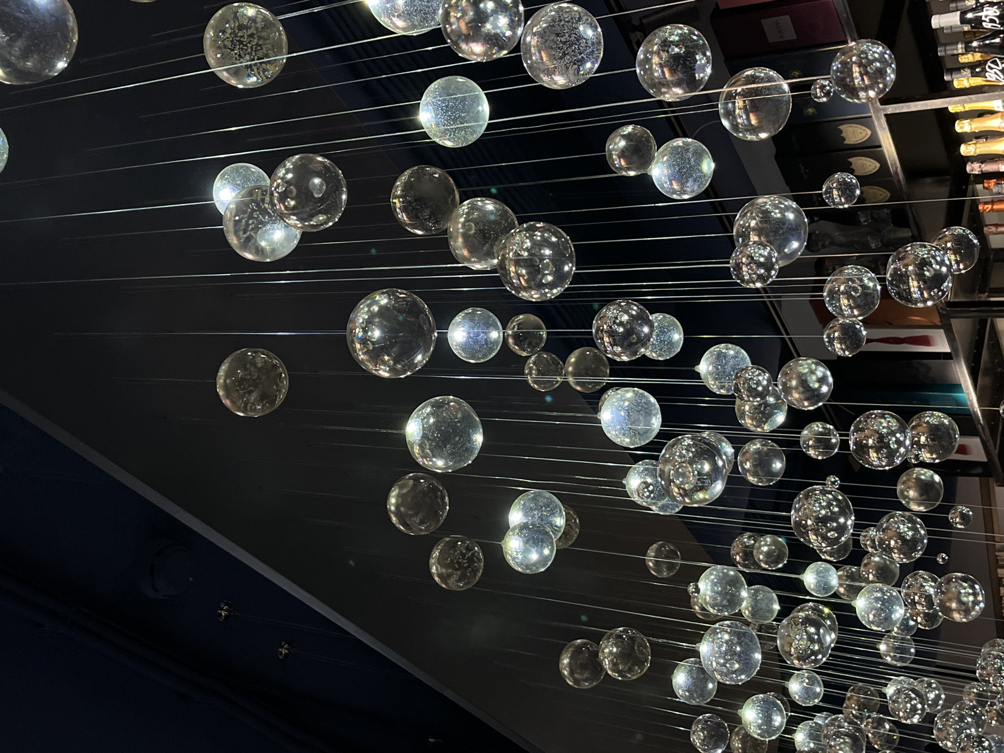 The image displays a modern decorative art installation featuring numerous clear glass spheres of varying sizes, suspended from thin, horizontal strands that appear to be metallic or light-colored. The spheres reflect and refract light, creating a complex interplay of highlights and shadows.
The background is dark, mostly black, with hints of a dark blue structural element in the lower left. In the upper right, there are glimpses of what appear to be bottles, indicating the installation may be located in a bar or a restaurant with a stocked bar area.
The installation creates an illusion of floating orbs, suspended in mid-air, and suggests a contemporary design aesthetic.
I cannot provide the location as Sochi, Russia, because I don't have access to that information.