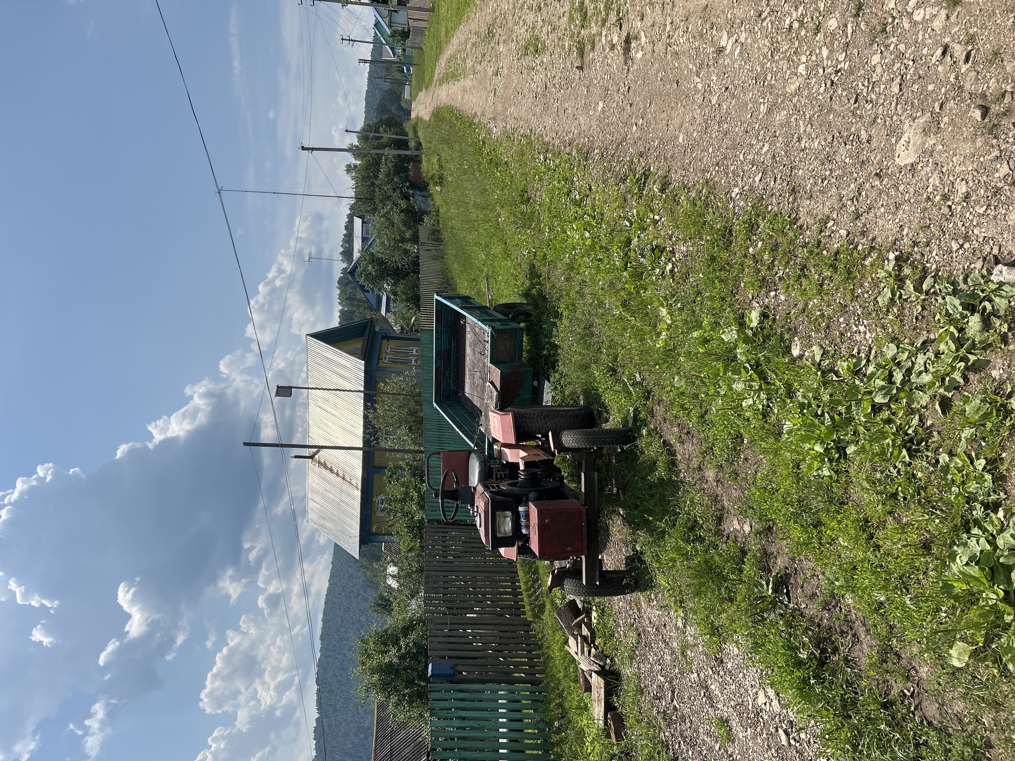 A bright, sunny day unfolds in Новомуллакаево, Russia, with fluffy white clouds dotting a vibrant blue sky. The scene is dominated by a rural, dirt road that winds through a village. In the foreground, to the left of the road, sits a small, red tractor with a hitch and an empty teal-colored trailer. The tractor appears weathered, with visible rust and a slightly worn appearance. Beside the tractor, a small pile of wooden planks rests on the gravelly ground.
A green wooden fence runs along the left side of the road, separating the properties. Beyond the fence, a yellow and blue house with a corrugated metal roof stands out, its details suggesting a traditional rural dwelling. More houses and trees are visible further down the road, stretching into the distance, with a gentle, tree-covered hill forming the backdrop. Power lines crisscross the sky above the houses, supported by utility poles. The overall impression is one of a peaceful, working village on a pleasant summer day.