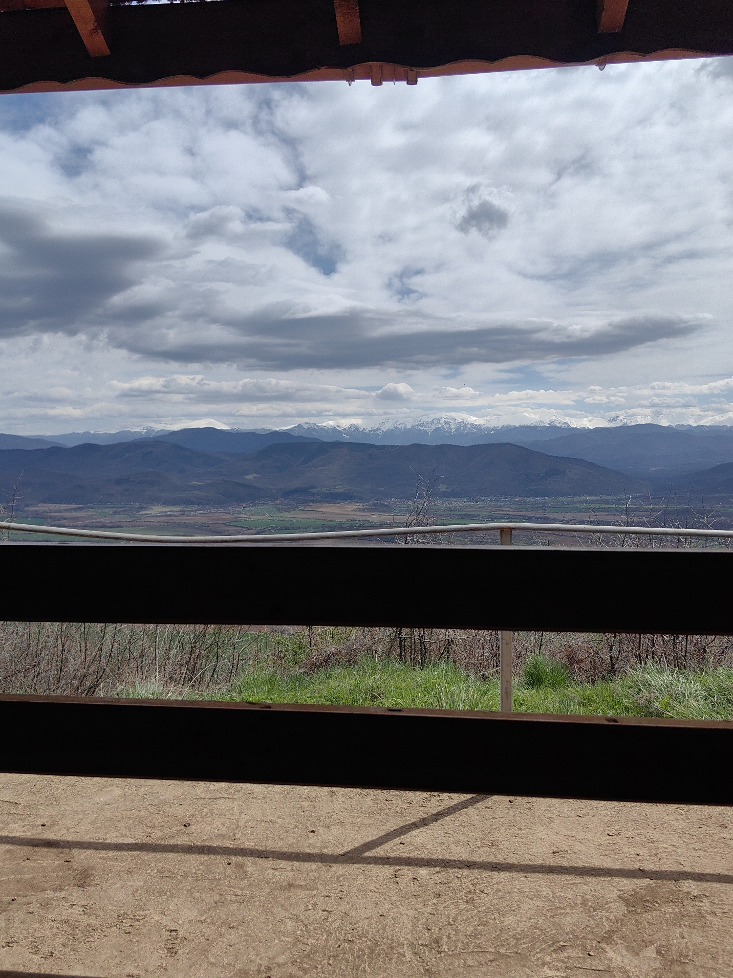 The image depicts a view of mountains and a valley from an elevated platform in Gumoshtnik, Bulgaria. The foreground features a weathered concrete floor and a wooden railing, casting shadows. Beyond the railing, there's a layer of vegetation including some grass and trees. In the middle ground, there's a wide valley with fields and scattered settlements. In the background, a mountain range with snow-capped peaks is visible under a sky filled with clouds.