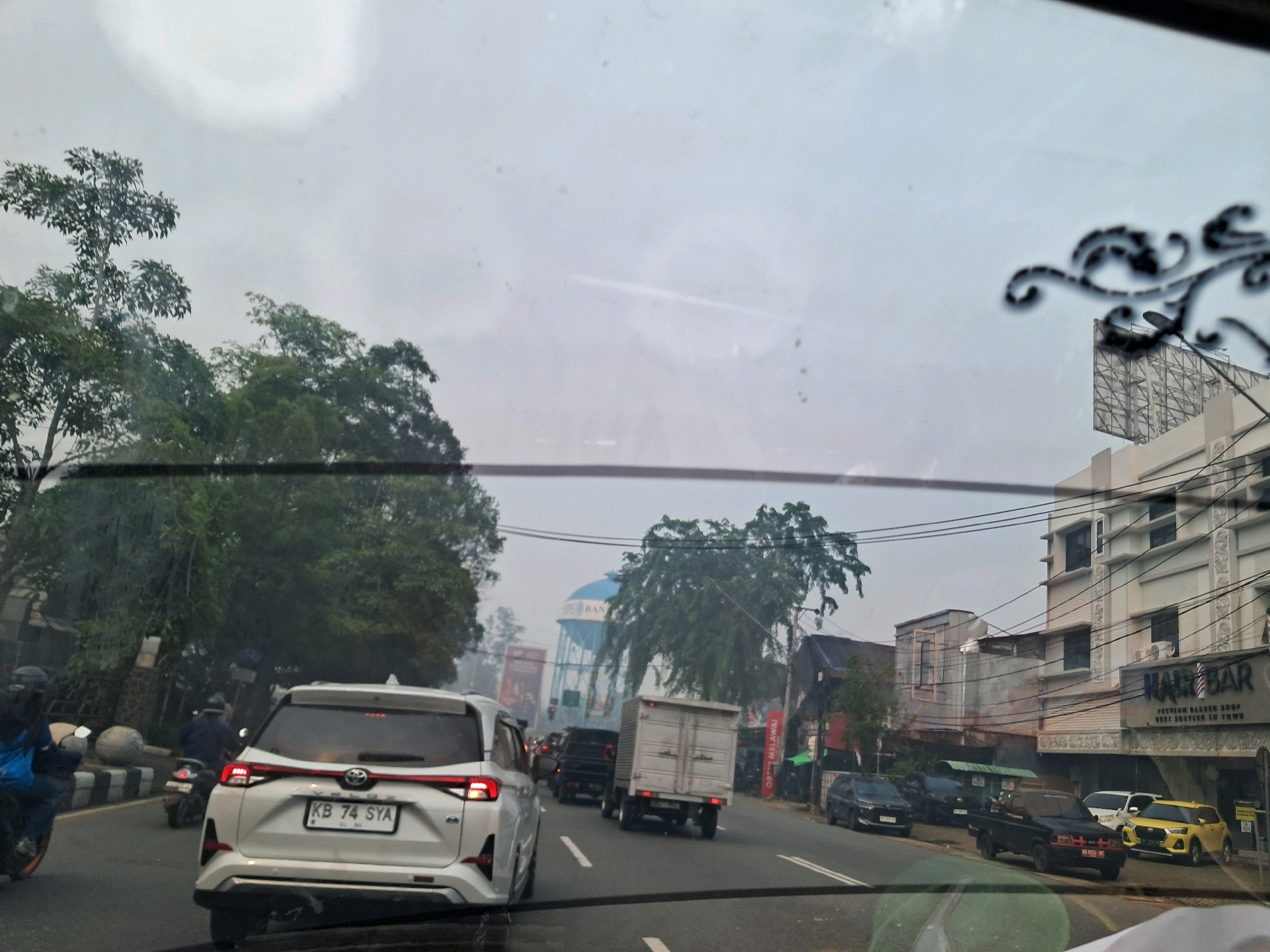 The image depicts a view from inside a vehicle traveling on a road in the city of Pontianak, Indonesia. The weather appears overcast or hazy. On the left side of the road, there are trees and some parked motorcycles, one of which is being ridden by a person wearing a blue backpack. Further down the road, a white SUV with a license plate reading "KB 74 SYA" is prominently in the foreground, followed by several other vehicles, including trucks and cars. On the right side of the road, there are buildings, including a multi-story structure with a sign that reads "HAIR BAR". There is also a water tower visible in the distance. The overall scene suggests a typical urban traffic environment with moderate congestion.