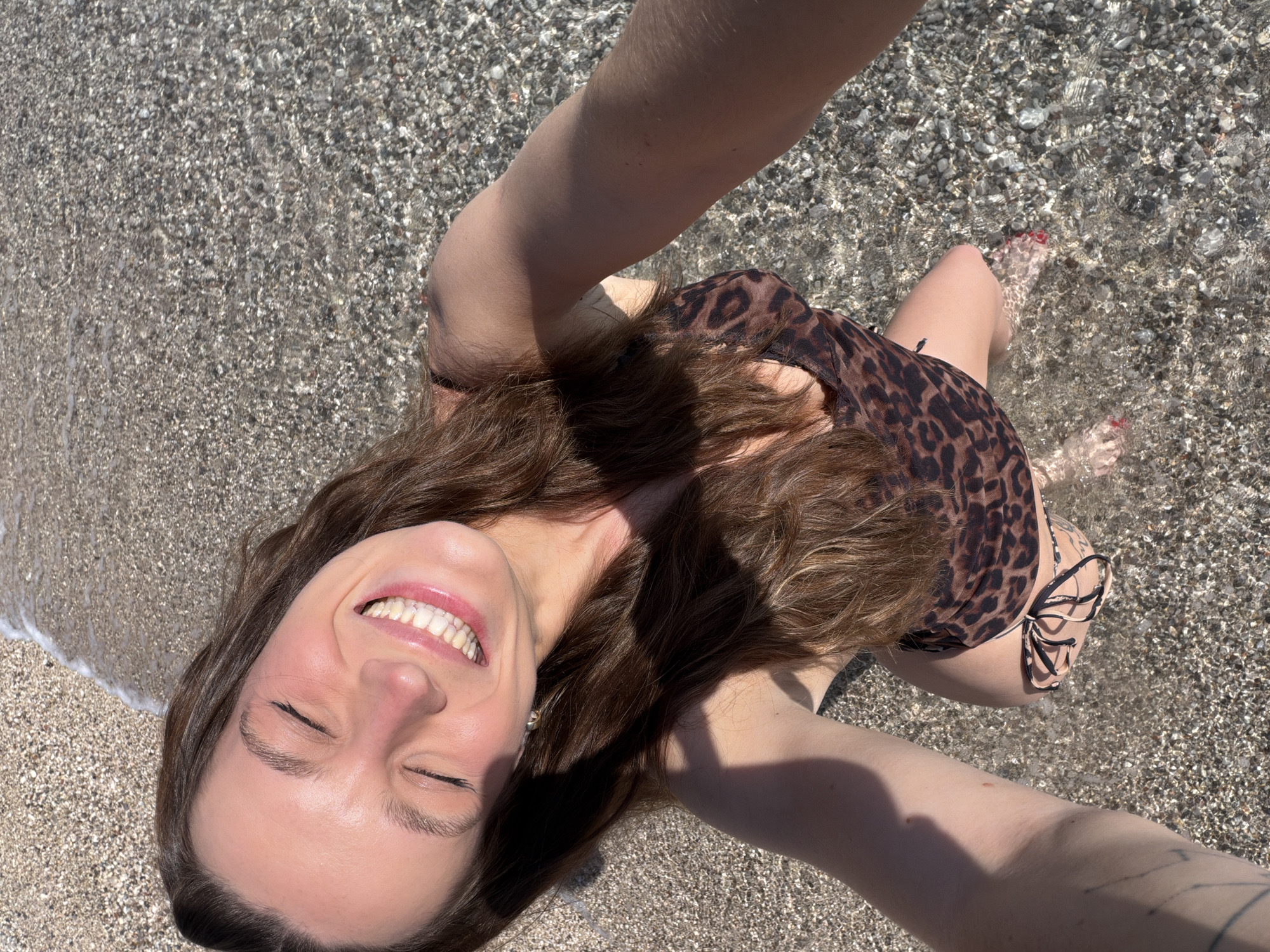 A medium, high-angle shot shows a smiling woman wearing a leopard-print swimsuit in the shallow water near a pebble beach. Her eyes are closed and her long brown hair is wet. Her arms are raised, presumably holding the camera. The water is clear and shallow, revealing the pebbles below. The scene is sunny. The location is Drobnići, Montenegro, as indicated by the pebble beaches characteristic of the area. The angle of the photo suggests the woman is lying on the beach, taking a selfie.