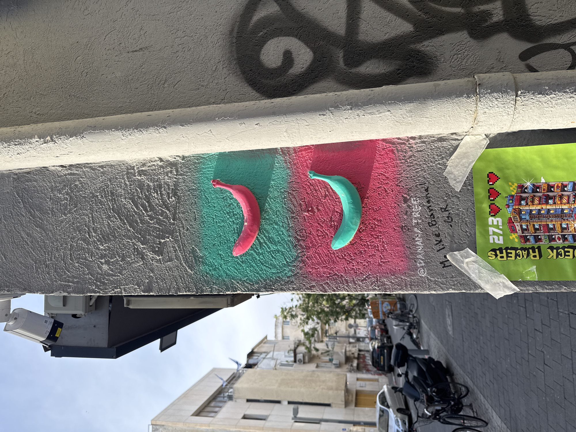 The image depicts an urban street scene in Tel Aviv, Israel, dominated by a light-colored, textured stucco wall under clear daylight. The wall features two distinct rectangular spray-painted sections: an upper teal-green area and a lower bright pink area. Affixed to these painted sections are two plastic banana sculptures: a pink banana within the teal-green area and a teal-green banana within the pink area. Below the painted sections, black handwritten text is visible, including "@BANANA_TREE" and "M. Live Banana! -G.R.". Below the text, a vibrant green poster is taped to the wall with white adhesive tape; it displays pixel-art graphics, including a multi-story building, the text "DECK RACERS," "27.3," and three red pixelated hearts. A vertical, off-white drainpipe runs along the right side of the wall, partially obscuring a large, dark grey shadow or graffiti tag.
Above and to the left of the wall, a white cylindrical security camera is mounted on a grey metal bracket extending from the building. The background showcases a typical Tel Aviv street: a grey paved sidewalk with several parked bicycles and a black scooter, leading to a street with parked vehicles including a silver sedan, a dark SUV, and other cars further down. Multi-story residential or mixed-use buildings with balconies, characteristic of the city's architecture, line the street, with a clear blue sky overhead. A street light pole and scattered trees are also visible in the distance.