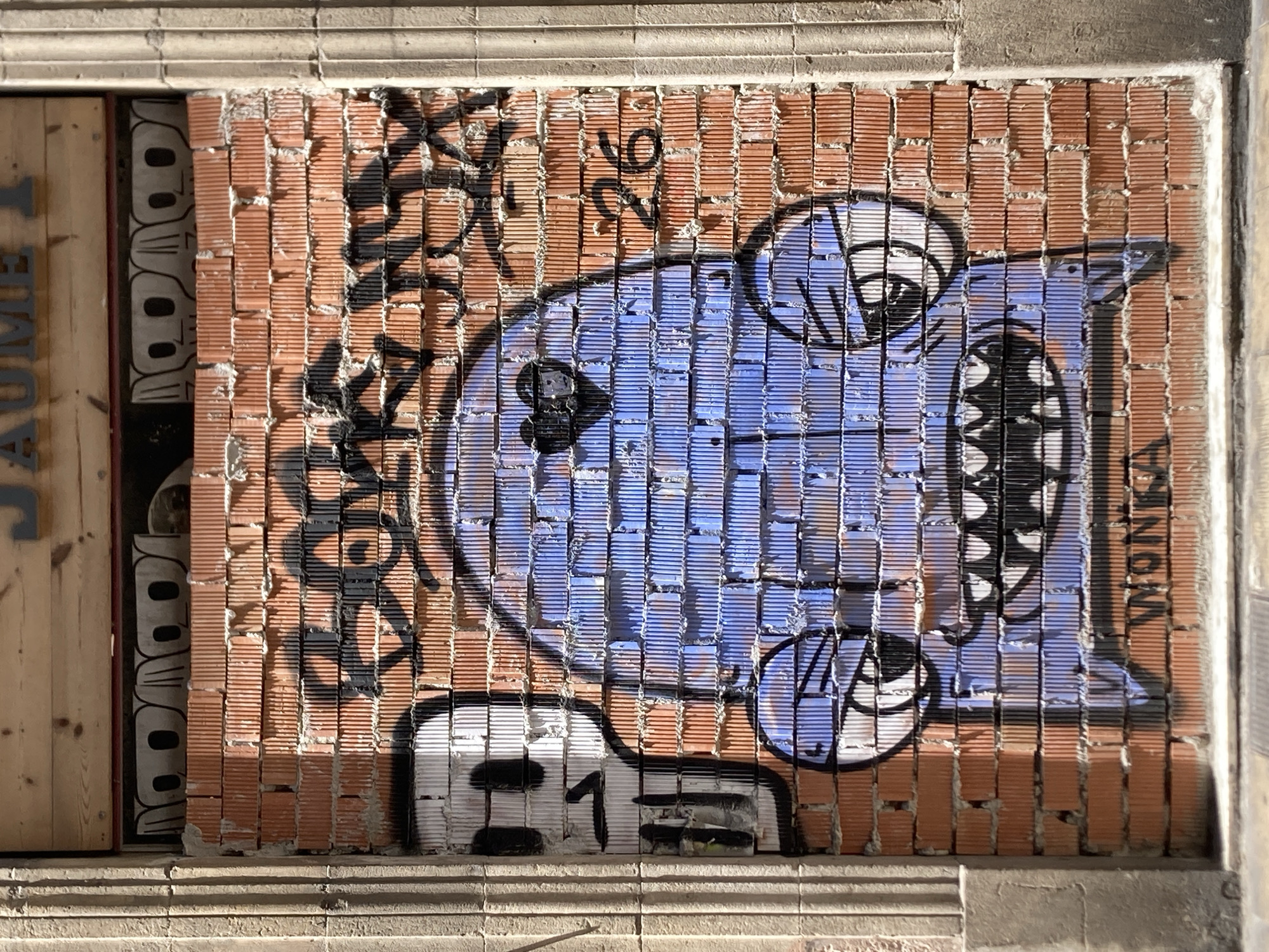 This image shows a graffiti art piece on a brick wall in Barcelona, Spain. The graffiti is vibrant and colorful, featuring a large, blue cartoon-like character with wide eyes and an open mouth filled with sharp teeth. The character is rendered in a somewhat abstract style, with bold black outlines. To the left of the character, there is black graffiti lettering, and to the right, the word "MONKA" is visible in black lettering. Below the main character, there are numbers painted in black and white, appearing to be "21". The brick wall itself is made of orange bricks, some of which are partially missing or broken, creating an uneven texture. The graffiti covers a significant portion of the wall. The lighting suggests it might be daytime, with shadows indicating the sun's position. The overall scene depicts an urban environment where street art is a prominent feature.