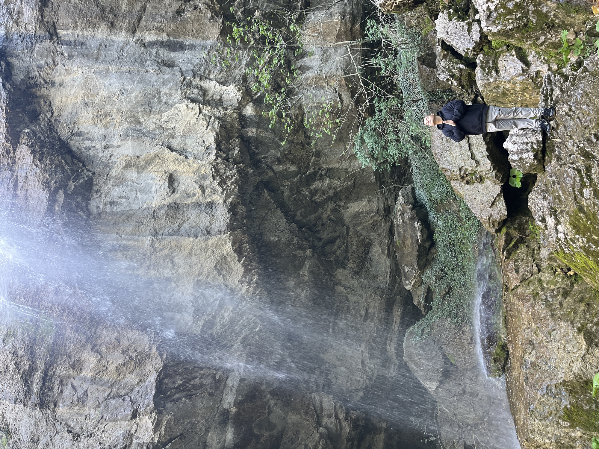 The image depicts a waterfall scene likely located in a rocky, natural environment. A cascade of water flows down a high, textured rock face, creating a misty effect in the foreground. To the right, a person stands atop a mossy, rugged rock outcrop adjacent to the waterfall. They appear to be an adult wearing dark attire, with their hands clasped together in front of them. Vines and greenery adorn the rock face in places, adding natural detail to the scene. Given the prompt, the scene may be Veselinovo, Bulgaria.