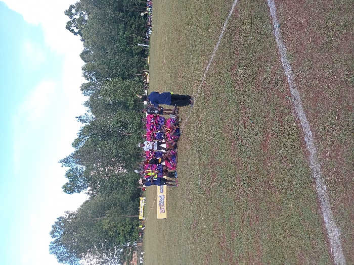 The image displays an outdoor scene on a sports field in Meru, Kenya. A group of approximately 15-20 individuals is clustered in the middle ground. The majority are dressed in matching pink and black athletic uniforms, with several members sitting or kneeling in the foreground while others stand behind them. A golden trophy is visible, held by an individual near the center of the standing group. An individual in a dark blue shirt and trousers stands in the right foreground, facing the assembled group. Another person in a white patterned shirt stands within the cluster. A yellow banner displaying "Betika!" in black lettering is laid horizontally on the ground directly behind the group. The field consists of sparse green grass intermixed with visible patches of reddish-brown earth. A white chalk line, indicative of a sports field boundary, is present in the bottom right corner. A dense line of tall, leafy green trees forms the background across the entire width of the image. The sky above is light blue with scattered white clouds, indicating daylight.
