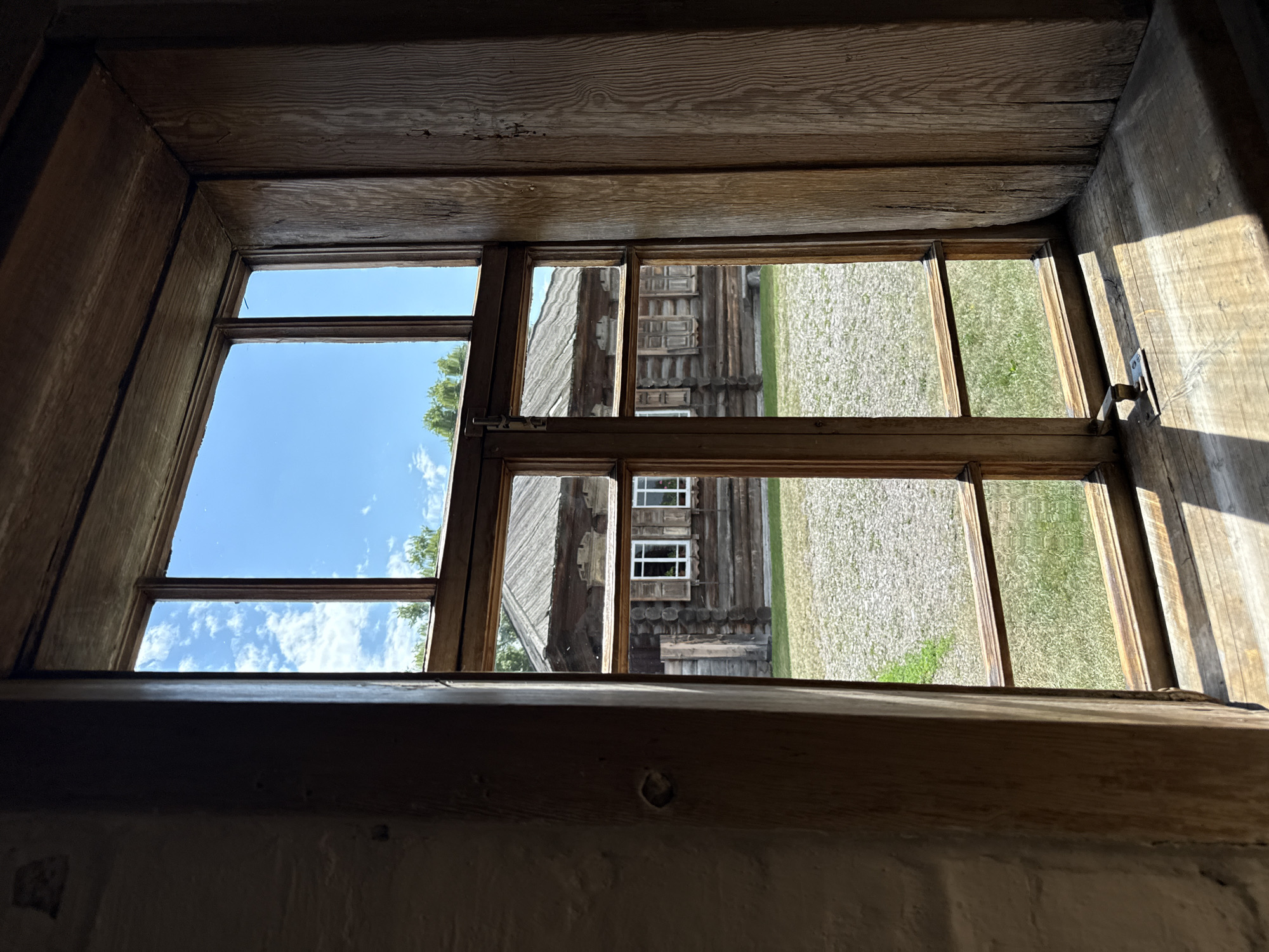 The image shows a view through a wooden-framed window onto an outdoor scene. The window frame itself is made of rough-hewn wood and is divided into several panes by horizontal and vertical wooden mullions. Sunlight streams in through the window, casting distinct shadows on the wooden windowsill and the floor beyond.
Through the window, a rustic wooden building with a pitched roof is visible. The building appears to be constructed from logs, a common architectural style in rural Russia. It has several windows with white panes and wooden shutters. The area directly in front of the building is covered with light-colored gravel and patches of green grass. In the background, trees are visible against a bright blue sky with scattered white clouds.
This scene is likely from the Shushenskoye museum-reserve, a historic site in Shushenskoye, Russia, dedicated to preserving the memory of Vladimir Lenin's exile there. The architectural style of the building and the rustic setting are consistent with the historical buildings found in such reserves. The image captures a moment of tranquility, with the warm sunlight illuminating the scene.