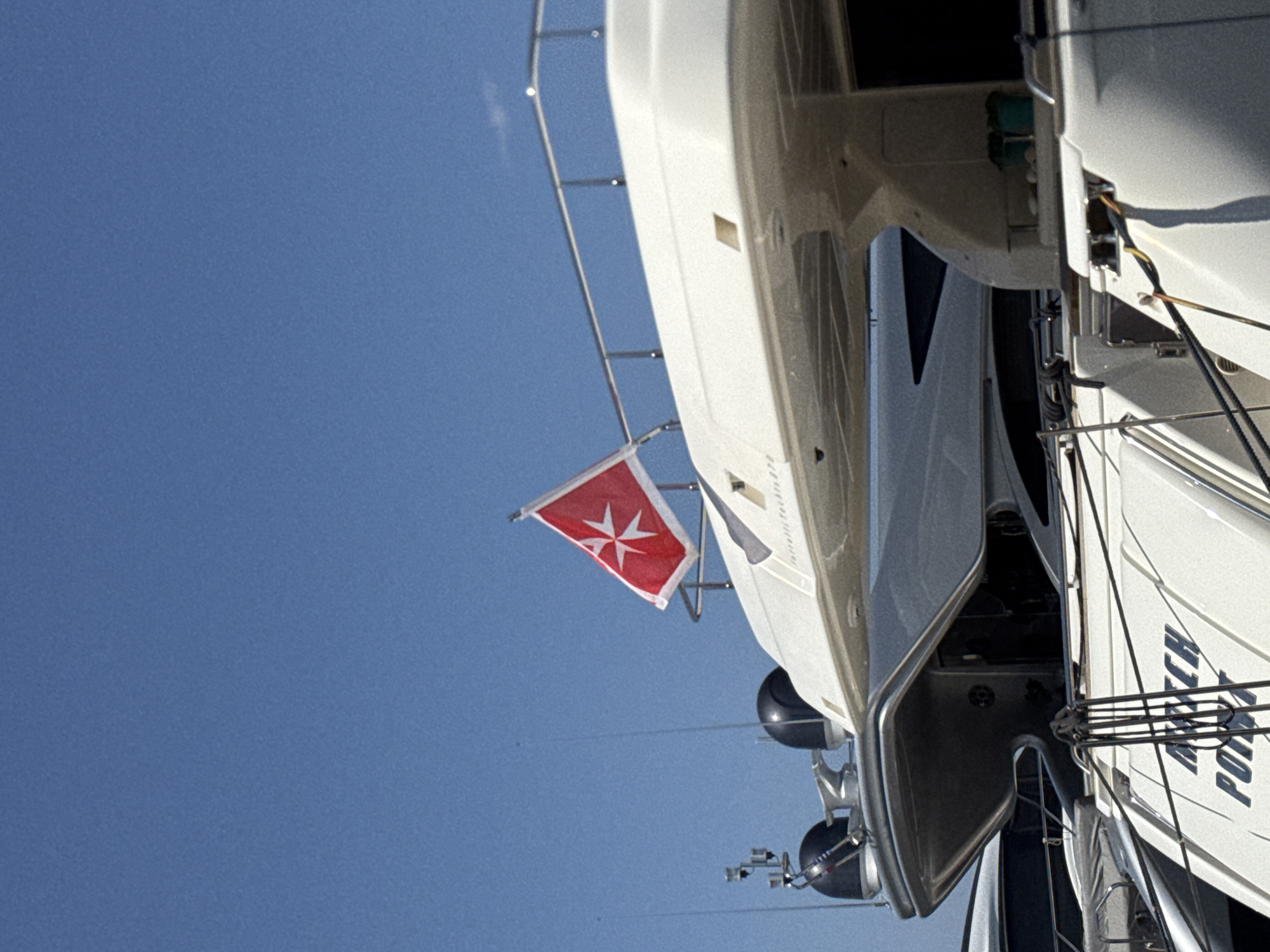 A close-up, low-angle exterior shot captures the upper decks of two docked yachts against a clear blue sky. A red flag with a white cross, likely the flag of the Order of Malta, flies from a railing on the larger yacht in the foreground. The yacht, painted white, has a sleek, modern design with dark-tinted windows visible on its upper structure. The name "FERRETTI YACHTS 870" is partially visible on the hull. In the background, the stern of another white yacht is visible, also docked. The lower portion of the foreground yacht displays the name "MATCH POINT" in black lettering. A few mooring lines are visible connecting the yachts. The scene appears to be a marina in Moschato, Greece, a port city southwest of Athens known for its shipbuilding and repair facilities. No people are visible in the frame.