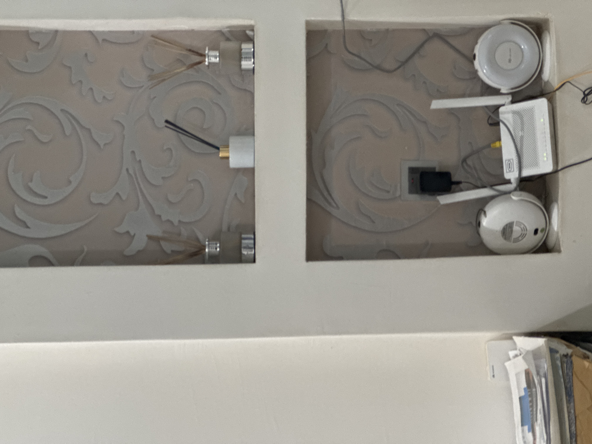 This is a medium-shot, eye-level, indoor photograph of a wall niche with two shelves. The wall is decorated with a light-colored, ornate wallpaper featuring swirling patterns.
On the top shelf, two glass diffuser bottles with reeds are visible. One bottle is white with a gold top, and the other is clear with a silver top.
The bottom shelf contains networking equipment. A white wireless router with two antennas is centrally placed. To the left and right of the router are two white circular devices, possibly Wi-Fi extenders or smart home devices. A black power adapter is plugged into a wall socket above the router. Cables are visible running from the equipment. To the far left, in the foreground, a stack of papers and envelopes is visible.
The image does not contain enough visual information to determine the specific location within Abuja, Nigeria.