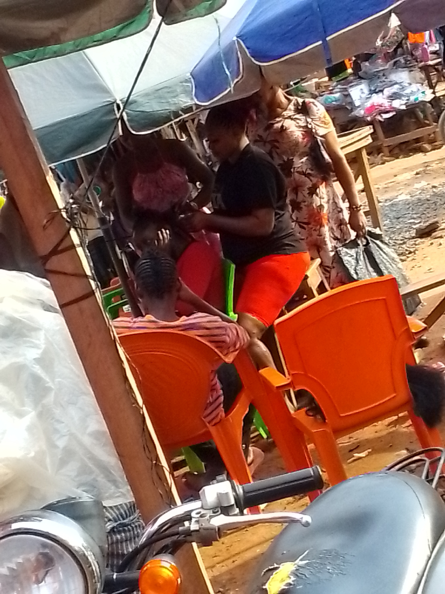 The scene is a marketplace setting in Orlu, Nigeria. People are present, with one woman in a floral dress holding a black plastic bag. Another woman is wearing a black shirt and red shorts. There are orange plastic chairs. The area is covered by umbrellas, and merchandise is displayed on tables. A motorcycle is in the foreground, with its handlebars and headlight visible. The ground appears to be dirt.