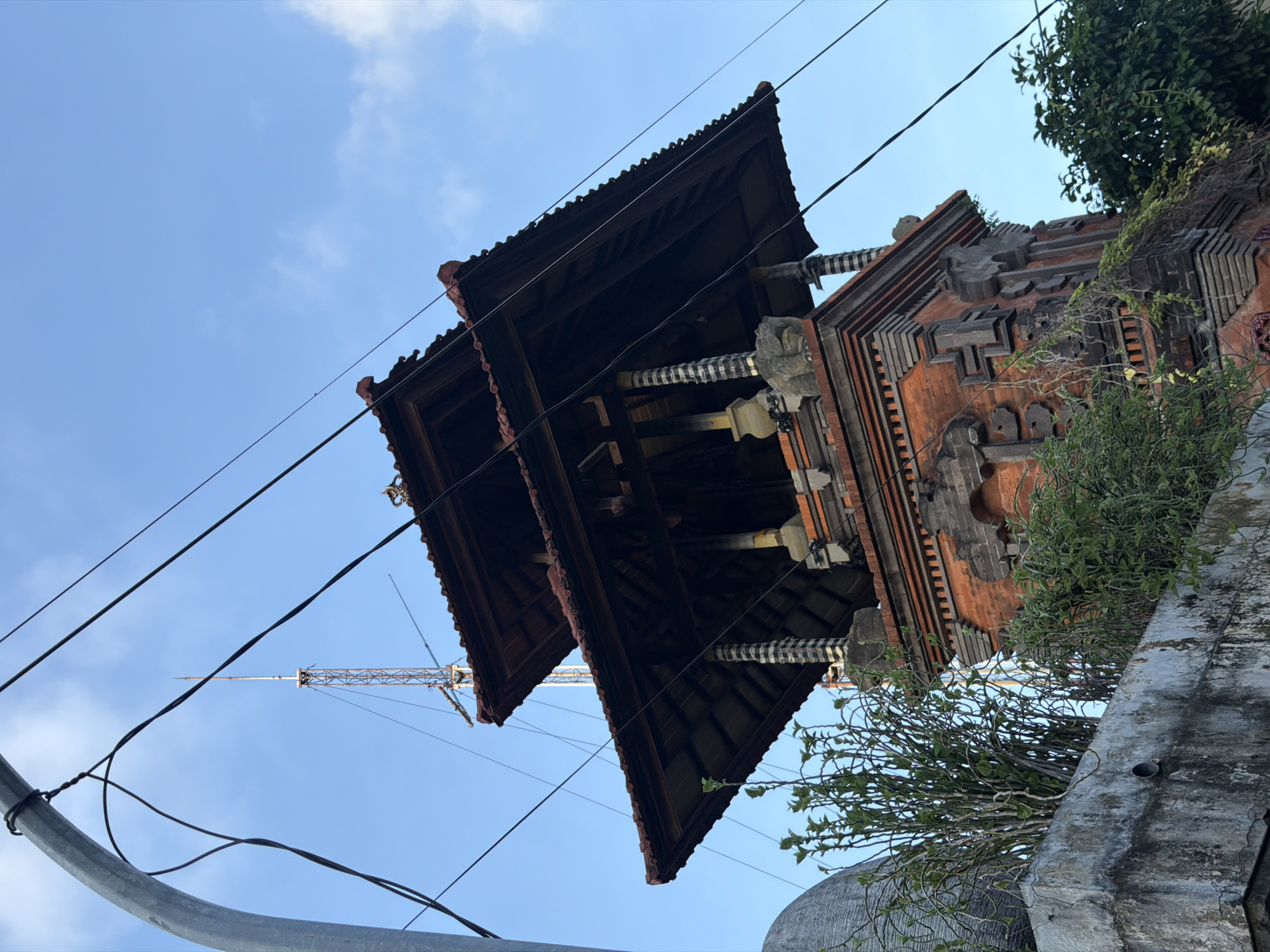 The media file depicts a low-angle shot of traditional Balinese architecture against a clear blue sky. The dominant subjects are the tiered roofs of a temple-like structure, constructed from dark wood with intricate detailing. The building is made of reddish-orange brickwork with ornate grey carvings and decorative elements. Lush green foliage spills over the edges of a concrete wall in the foreground, partially obscuring the structure. Several power lines crisscross the frame, adding a modern contrast to the ancient architecture. In the background, a tall radio tower or construction crane is visible against the sky, suggesting an urban or semi-urban setting. The lighting indicates it is daytime, likely late afternoon given the warm light and soft shadows. The scene conveys a sense of cultural heritage juxtaposed with contemporary infrastructure. No people are visible in the frame. There is no visible text. The location is Kuta, Indonesia.