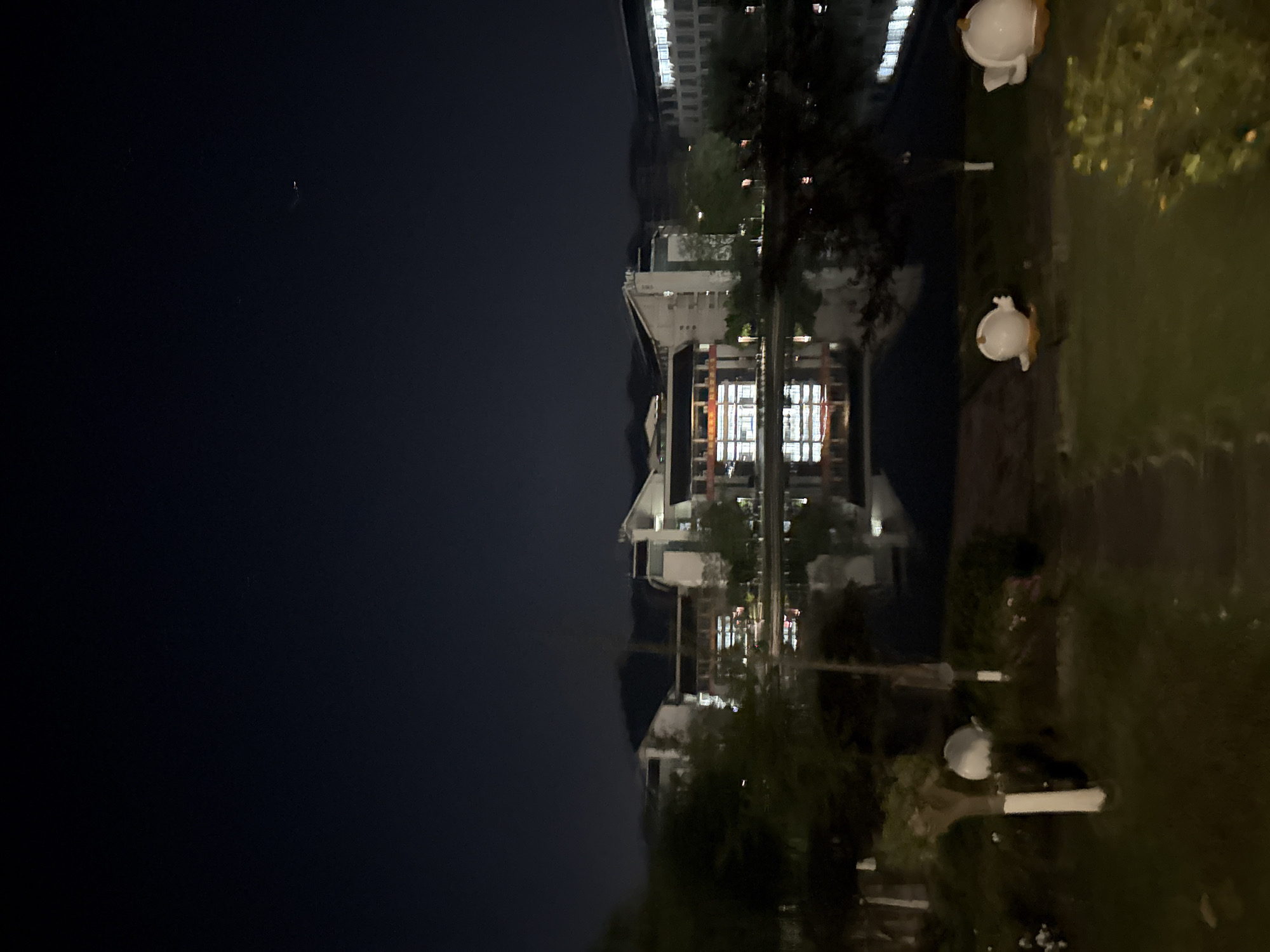 The image depicts a nighttime scene in Jiangjin, China, featuring buildings reflected in a body of water. The sky is dark, with faint stars visible. Structures, presumably residential or commercial, are illuminated with interior lights, their reflections visible on the water's surface. Landscaping includes trees and greenery along the water's edge. White, round decorative features are sparsely placed on the grassy bank.