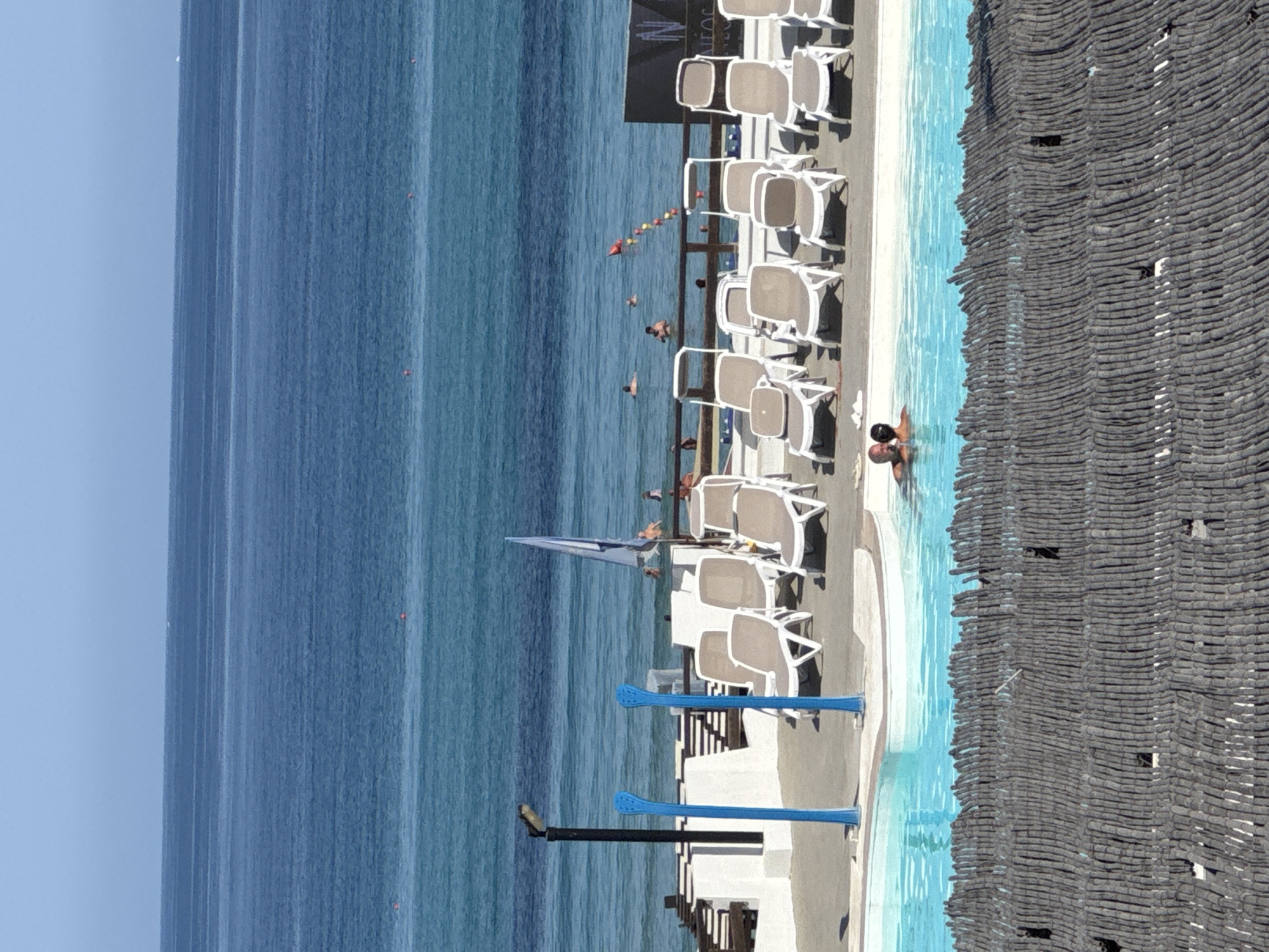 This is an outdoor shot of a swimming pool area next to the ocean. The pool is in the foreground, with bright blue water. There are two people in the pool, embracing. Beyond the pool, there is a wooden fence and then a paved area with rows of lounge chairs. In the background, the ocean stretches to the horizon, with several small buoys visible on the surface. A flag pole with a flag is also visible. The scene appears to be a resort or hotel in Isola delle Femmine, Italy, given the characteristic blue Mediterranean Sea and the architectural style of the buildings. The weather is sunny and clear.