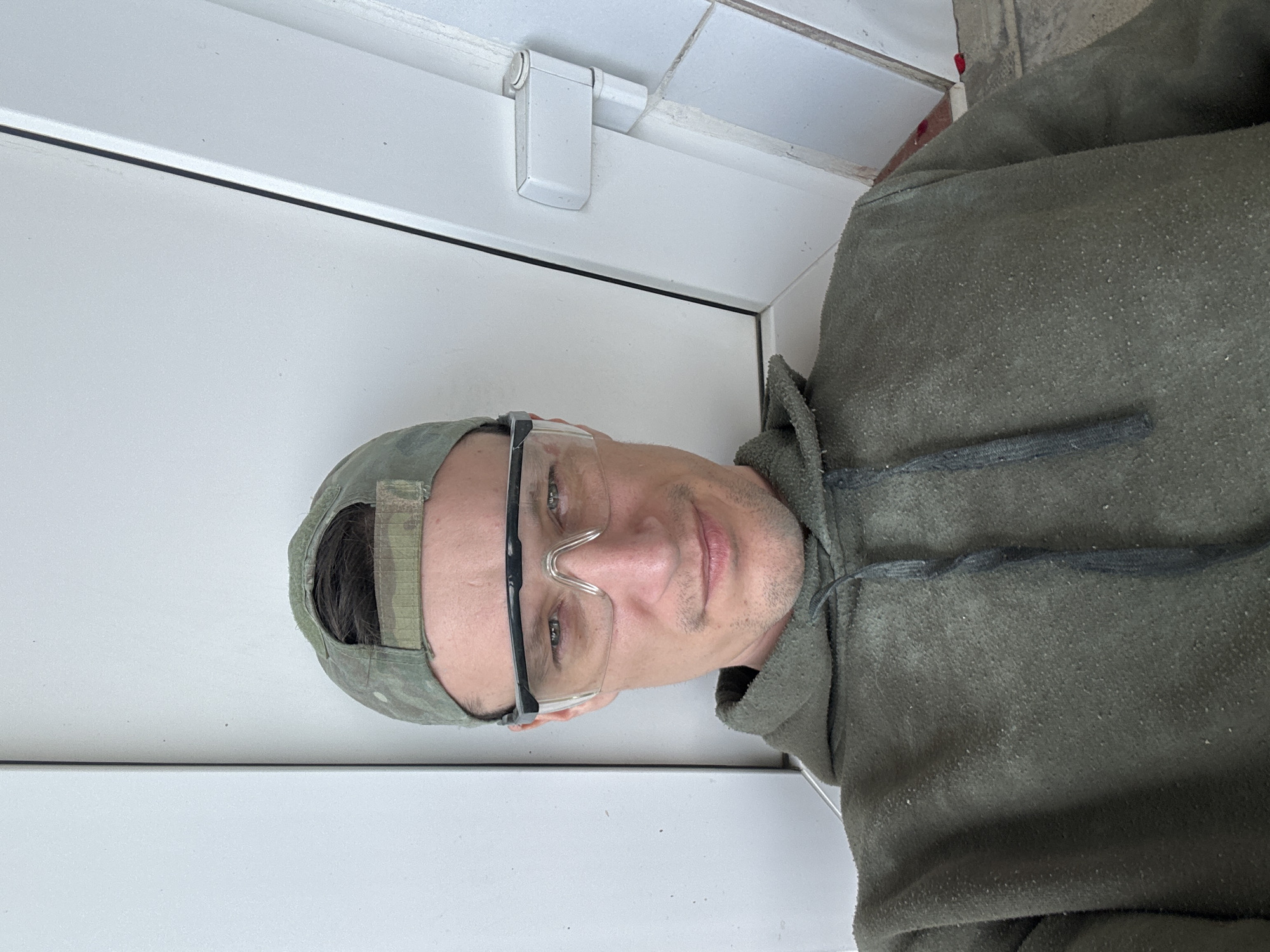 A man wearing a camouflage baseball cap, clear safety glasses, and a green hooded sweatshirt is sitting and looking at the camera. The background appears to be an interior space with white walls and a white door frame with a white hinge visible. The man's skin is fair, and he has short brown hair and stubble on his face. He is smiling slightly. There is no indication of the specific location within the city of Makiv, Ukraine.