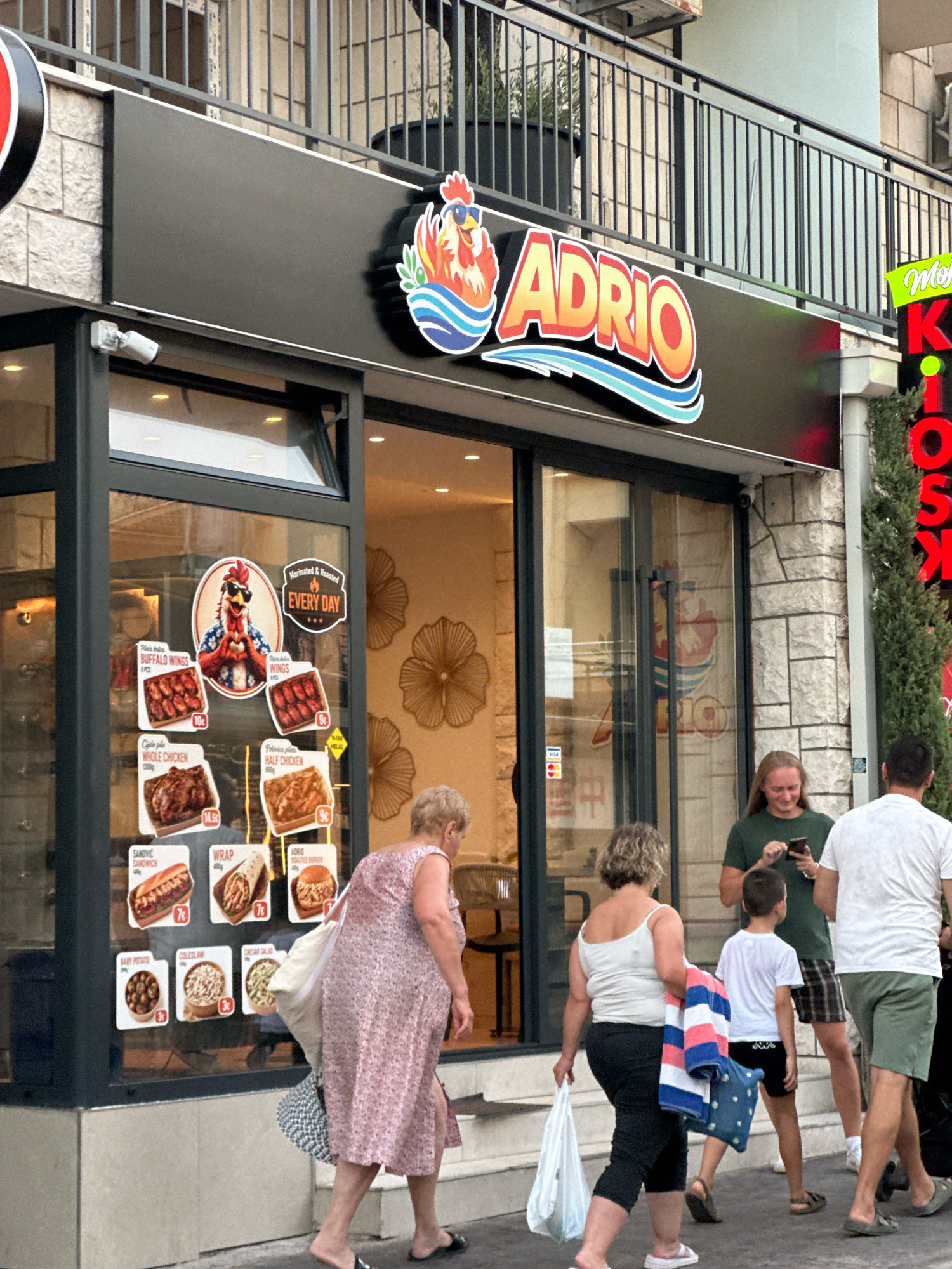 A group of people are walking past a fast-food restaurant called "ADRIO" in Budva, Montenegro. The restaurant has a large sign with a cartoon chicken wearing sunglasses. The window displays pictures of chicken meals, sandwiches, and salads. The people in the image include a woman in a pink floral dress carrying a tote bag, a woman in a white tank top and black pants carrying a plastic bag and a towel, a boy in a white t-shirt and black shorts, and two men. One man is wearing a green t-shirt and plaid shorts, looking at his phone. The other man is wearing a white t-shirt and green shorts. The restaurant is located on a street with stone buildings and a balcony above. The overall scene appears to be a daytime street view.