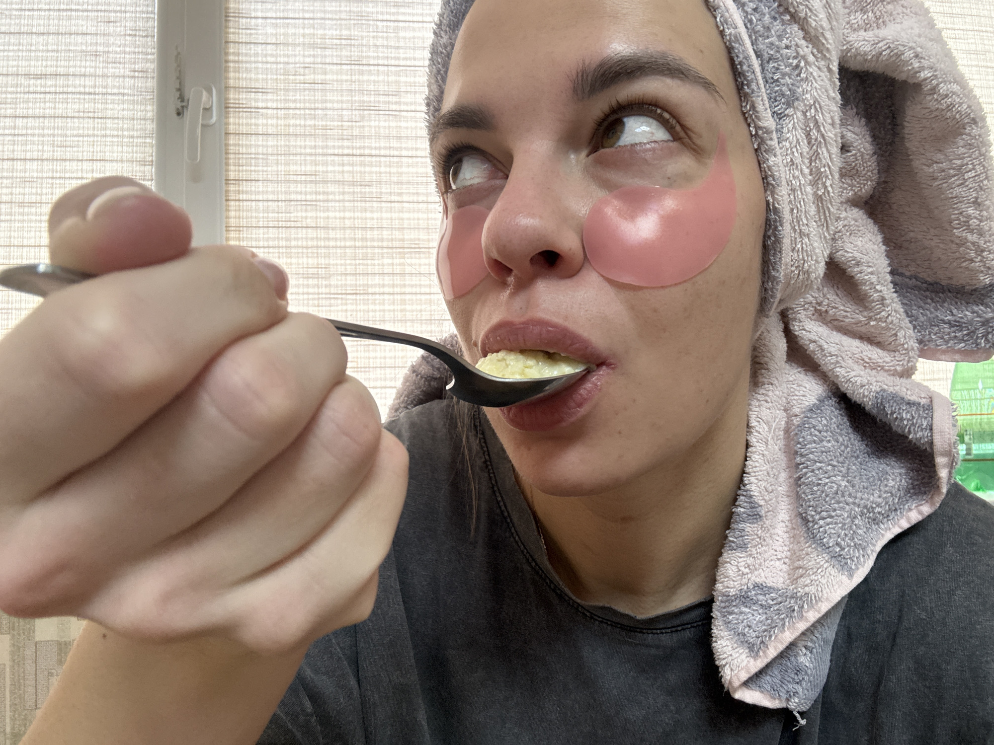 A person with pink under-eye patches and a towel wrapped around their head is eating from a spoon. The person is wearing a gray t-shirt. The background shows a window with blinds. There is no evidence to suggest that this image was taken in Ust-Ilimsk, Russia.