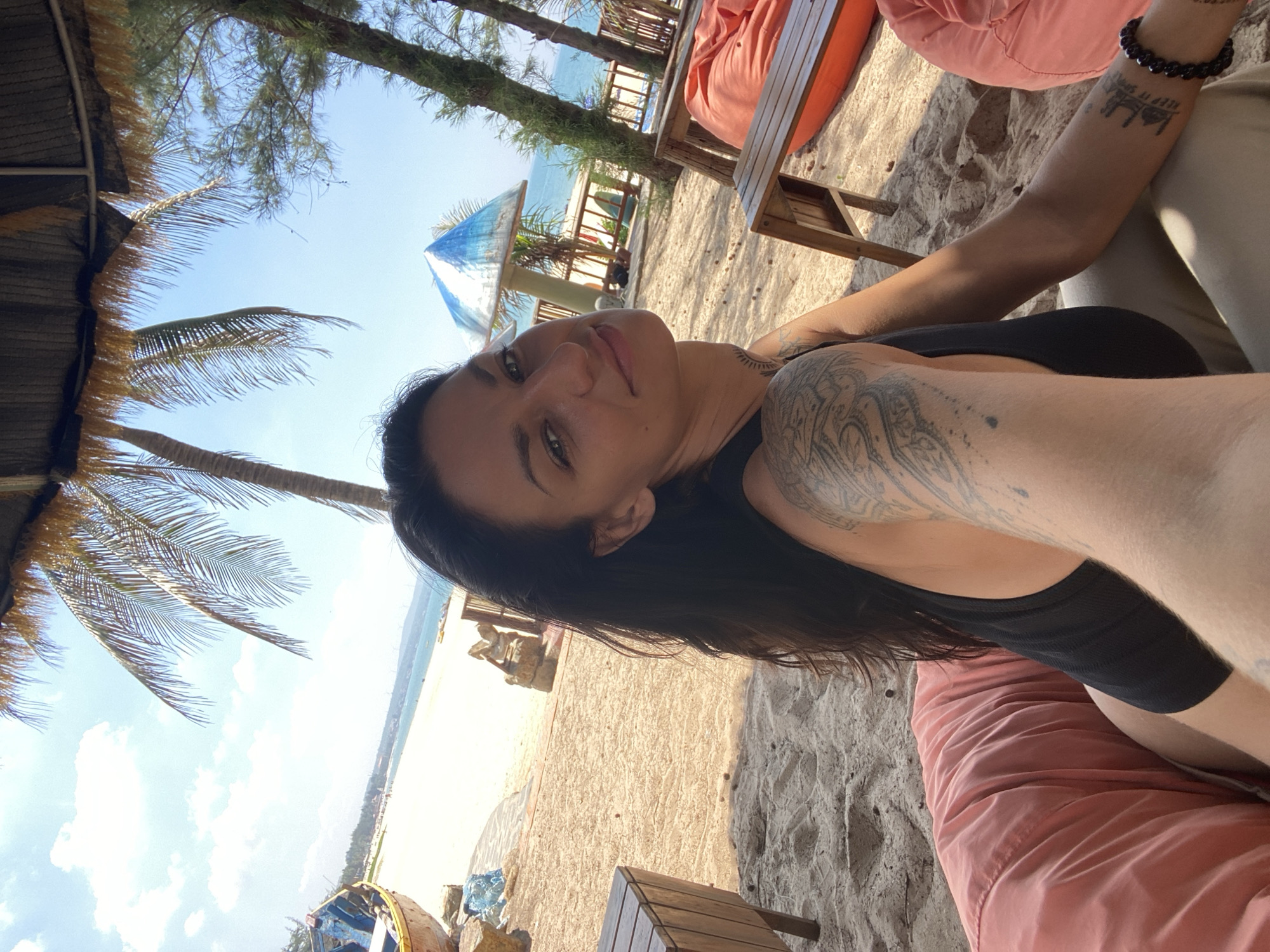 A woman with dark hair and tattoos on her arm is seated on a peach-colored beanbag chair on a sandy beach. She is wearing a black tank top and beige pants. In the background, palm trees and a thatched roof are visible, as well as the ocean and a blue and white umbrella-shaped structure. The scene appears to be located in Phu Thuy Ward, Vietnam.