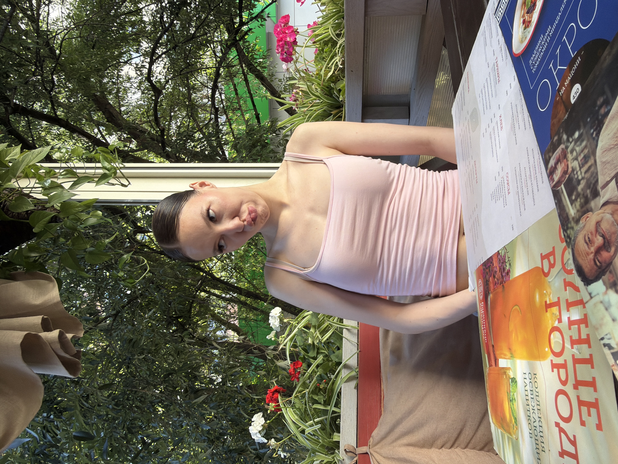 In this outdoor shot, a young woman with her hair pulled back in a bun is seated at a table. She wears a pale pink tank top and makes a pouty face towards the camera. The table is covered with menus. In the background, a planter box with red and white flowers is visible, and lush green trees provide shade overhead.
