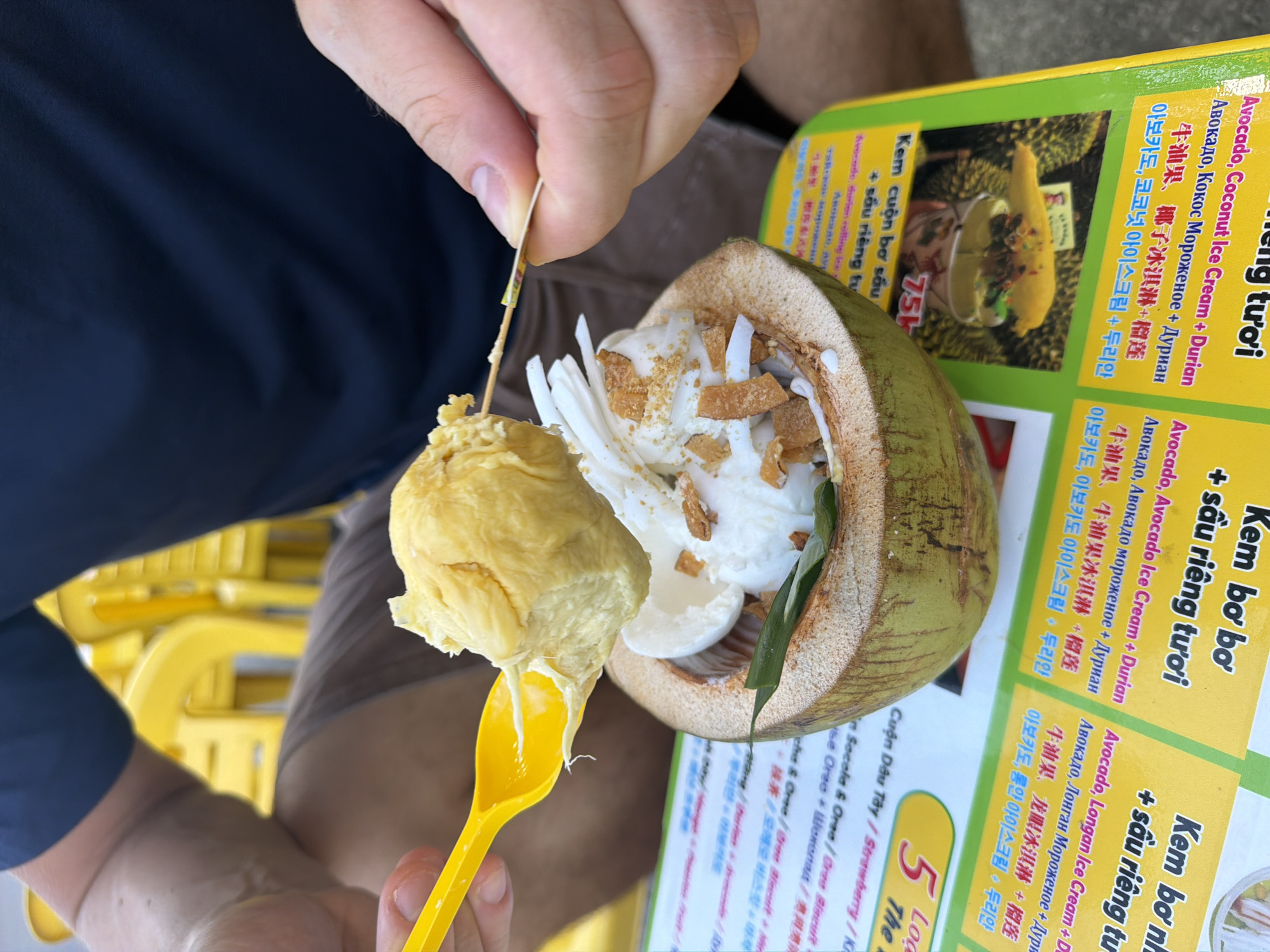 A close-up, eye-level shot shows a person holding a yellow plastic spoon with a piece of durian fruit in Mũi Né, Vietnam. The durian is being lifted from a half coconut shell filled with white ice cream, shredded coconut, and small brown crispy pieces, likely fried coconut flakes. The coconut shell rests on a laminated menu that is partially visible. The menu features text in multiple languages, including Vietnamese, Russian, and Korean, with prices displayed in Vietnamese Dong (75k). In the background, yellow plastic chairs are visible, suggesting an outdoor seating area. The person's hands, one holding the spoon and the other a toothpick, are in focus in the foreground. The overall scene depicts a refreshing dessert being enjoyed in a casual setting.