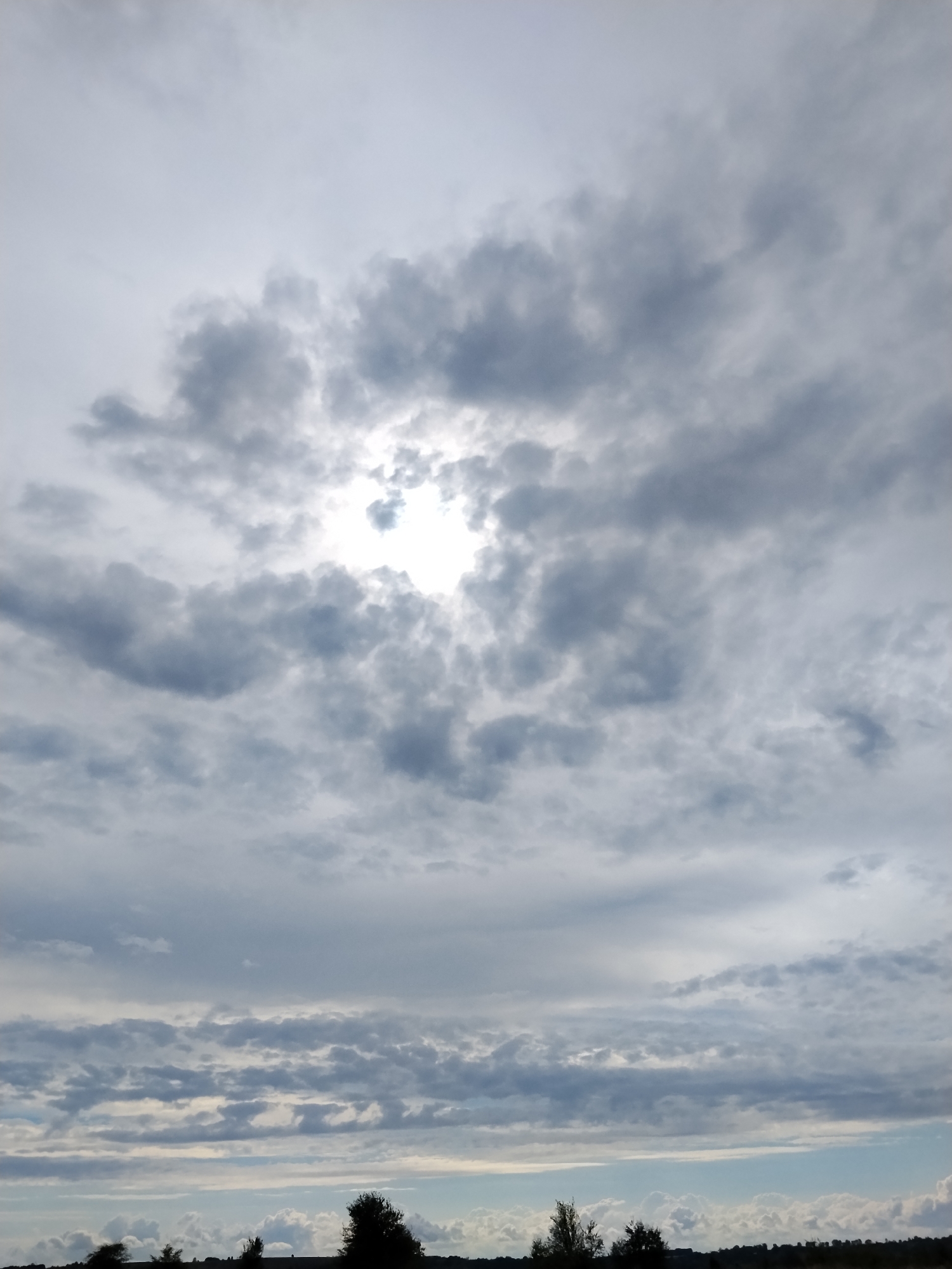 The image depicts a cloudy sky over a landscape. The sky is filled with various cloud formations, ranging from light and wispy to dark and dense. The sun is partially visible through a break in the clouds, creating a bright spot in the upper center of the frame. Below the sky, a silhouetted landscape is visible, consisting of a horizon line with scattered trees and possibly some undulating terrain. The overall impression is of a somewhat overcast day with patches of sunlight. The location is identified as Ustyvytsya, Ukraine.