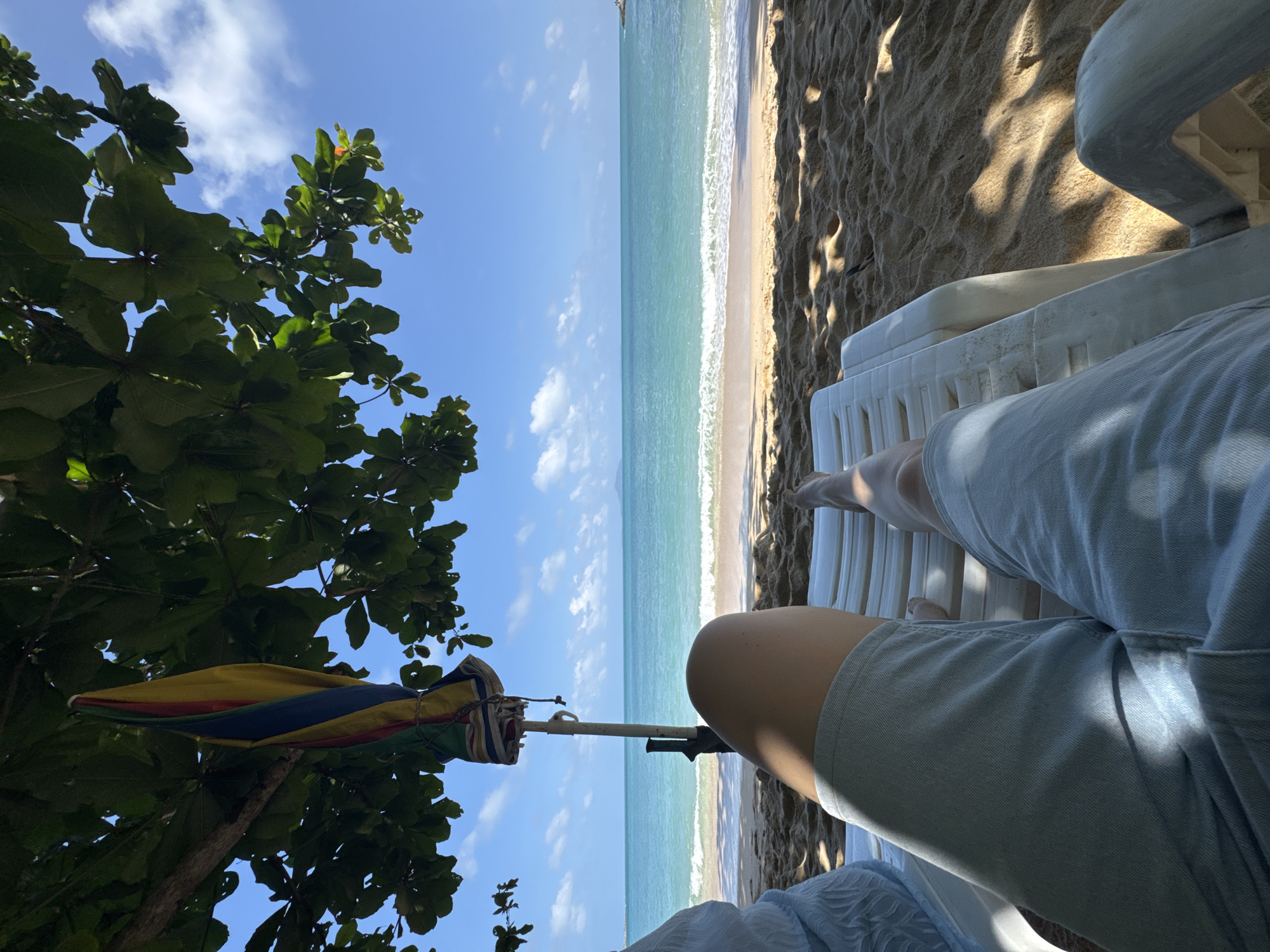 A first-person perspective shot captures a person relaxing on a white plastic beach chair on a sandy beach in Rawai, Thailand. The individual, wearing light blue denim shorts, is seated and facing the ocean. Their legs are visible, one bent and the other extended. To the left, a blue textile is partially visible, draped over what appears to be another chair or part of the seating.
Above the person, a portion of a large green tree with broad leaves extends into the frame, casting dappled shadows on the sand and the beach chair. Hanging from a branch of this tree is a closed beach umbrella with vertical stripes of yellow, red, and blue. A pole, likely supporting the umbrella, is visible next to the seated person.
The background is dominated by the turquoise ocean with gentle waves lapping at the shore. The water transitions to a lighter shade near the sandy beach, which is marked by wet sand and the remnants of small waves. Beyond the water, a faint landmass is visible on the horizon under a bright blue sky dotted with fluffy white clouds. The overall scene conveys a sense of peaceful relaxation on a sunny day.