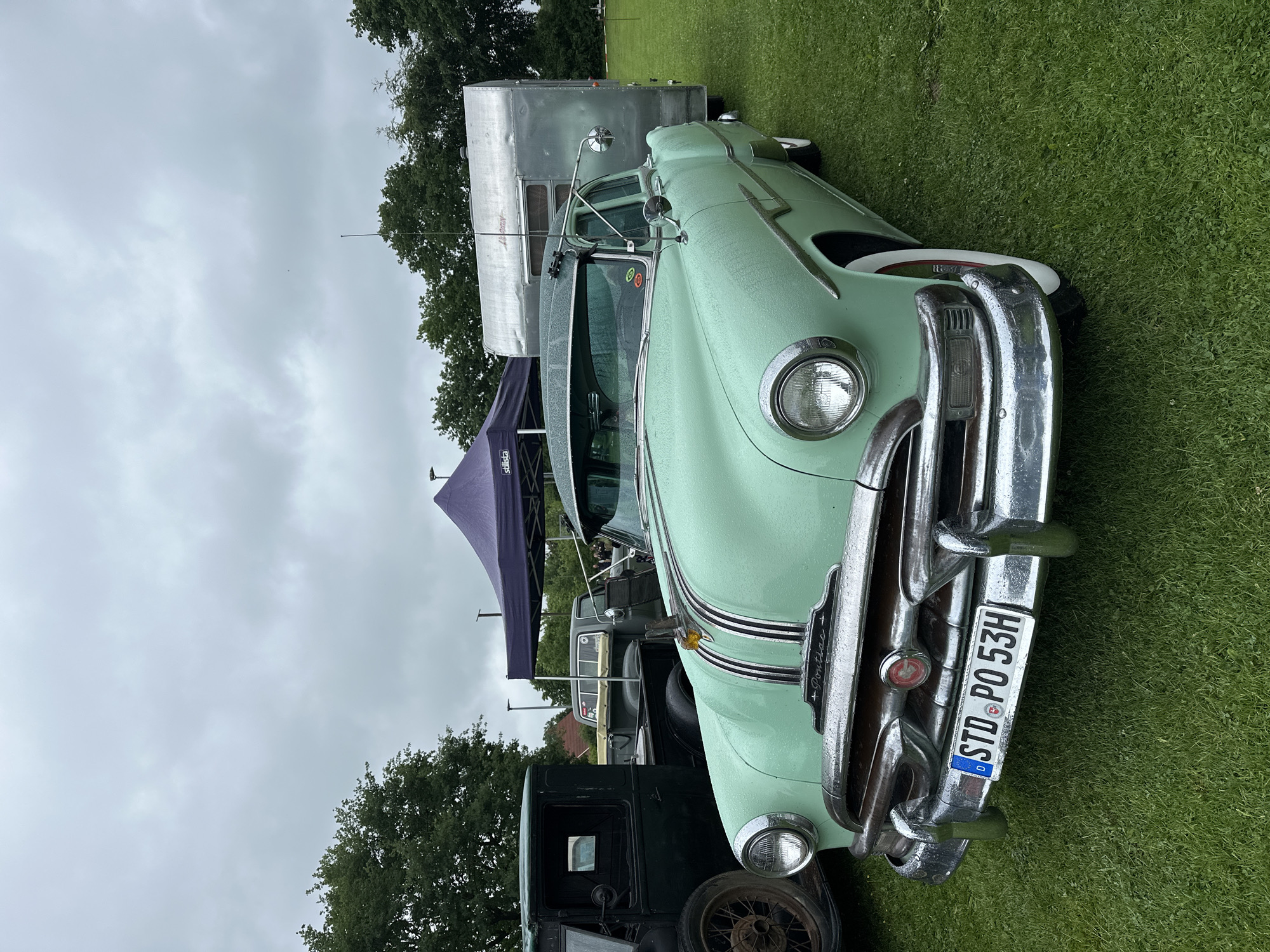 An outdoor scene in Deinste, Germany, depicts a light green, 1950s-era Pontiac sedan parked on a grassy field under an overcast sky. The vehicle is angled towards the viewer, slightly to the right, showcasing its chrome bumper, grille, and round headlights. Water droplets adhere to the car's surfaces, including the paintwork and chrome, indicating recent precipitation. A German license plate, "STD PO 53H," is mounted on the front bumper. The car features a hood ornament, an external sun visor above the windshield, and an auxiliary rearview mirror on the driver's side. White sidewall tires are visible. To the left, a portion of an older, dark green truck is visible, including its front wheel and cabin side. To the right, the Pontiac is hitched to a vintage, silver-colored travel trailer with a rounded design. In the background, a purple pop-up tent, partially displaying "Stilista," is erected. The perimeter features lush green trees. The ground is covered in green grass. No people are visible.