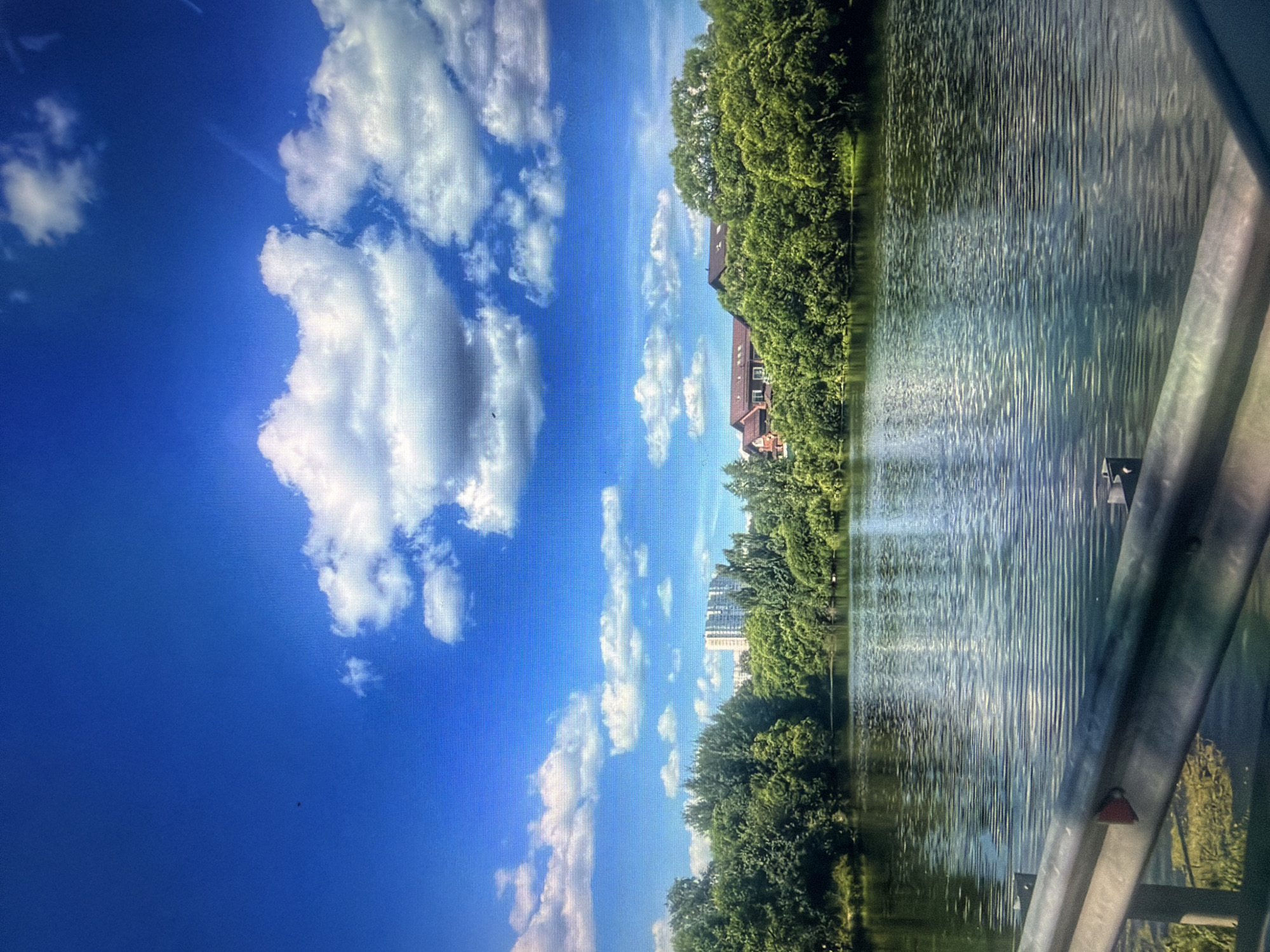 A wide-angle, slightly distorted, eye-level shot captures a serene body of water reflecting the bright blue sky dotted with fluffy white clouds. A metal guardrail in the foreground runs horizontally across the bottom of the frame, partially obscuring the view. The middle ground features a lush, dense line of green trees along the opposite bank of the water. Beyond the trees, a modern, light-colored multi-story apartment building is visible in the distance, suggesting an urban or suburban setting. To the right, nestled amongst the trees, are residential buildings with reddish-brown roofs. The overall impression is of a tranquil natural scene with man-made structures in the background. This scene is located in the city of Kotelniki, Russia.