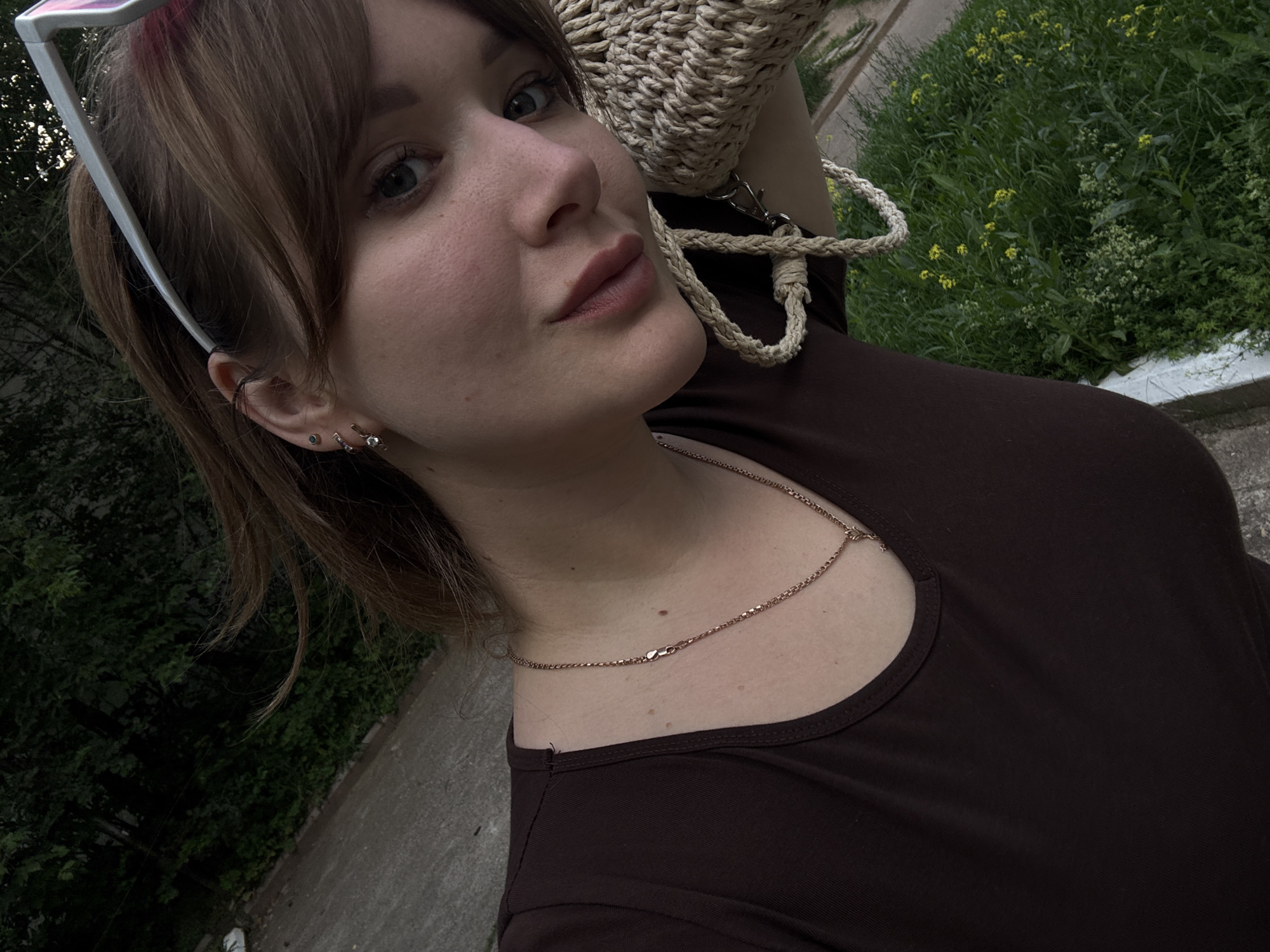 The image is a close-up selfie of a young woman with shoulder-length brown hair. She is wearing a dark brown t-shirt and a delicate gold necklace. She has multiple piercings in her left ear, including a small hoop and studs. She is also wearing white-rimmed sunglasses perched on her head, revealing pink-tinted lenses. A woven straw bag with a thick braided strap is slung over her shoulder, partially obscuring the right side of her face. Her makeup includes subtle blush and lip color.
The background consists of lush green foliage and trees, with a paved surface visible in the lower left and upper portions of the image, suggesting an outdoor urban or park setting. The lighting indicates it is daytime, possibly late afternoon due to the soft light. There is no specific identifying information to pinpoint the location to Chaikovsky, Russia.